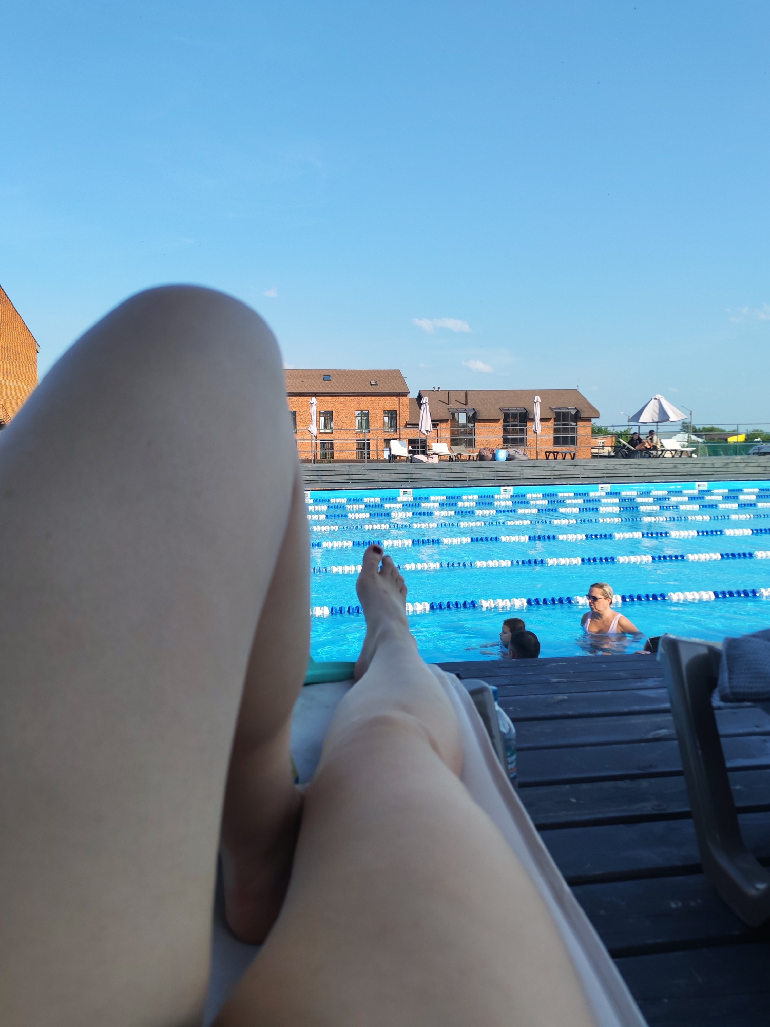 The image depicts a sunny day at an outdoor swimming pool in Pirogovo, Russia. The foreground is dominated by the legs of a person lying on a lounge chair, facing away from the camera. The lounge chair is dark wood, and a partially visible bottle of water sits next to it. Beyond the lounge chair, a blue swimming pool with lane markers stretches out. In the pool, several people are visible. A woman in a white swimsuit and sunglasses is standing in the water, with two children near her. Further back, other swimmers and people relaxing by the pool are discernible.
Behind the pool, there are brick buildings with multiple windows and arched openings, suggesting a resort or residential complex. White umbrellas are positioned on a deck area next to the pool. The sky is clear and bright blue, indicative of fair weather. The overall scene conveys a relaxed atmosphere of a summer day spent by the pool. The time of day appears to be late afternoon, given the angle of the sunlight. No visible text is present in the image.