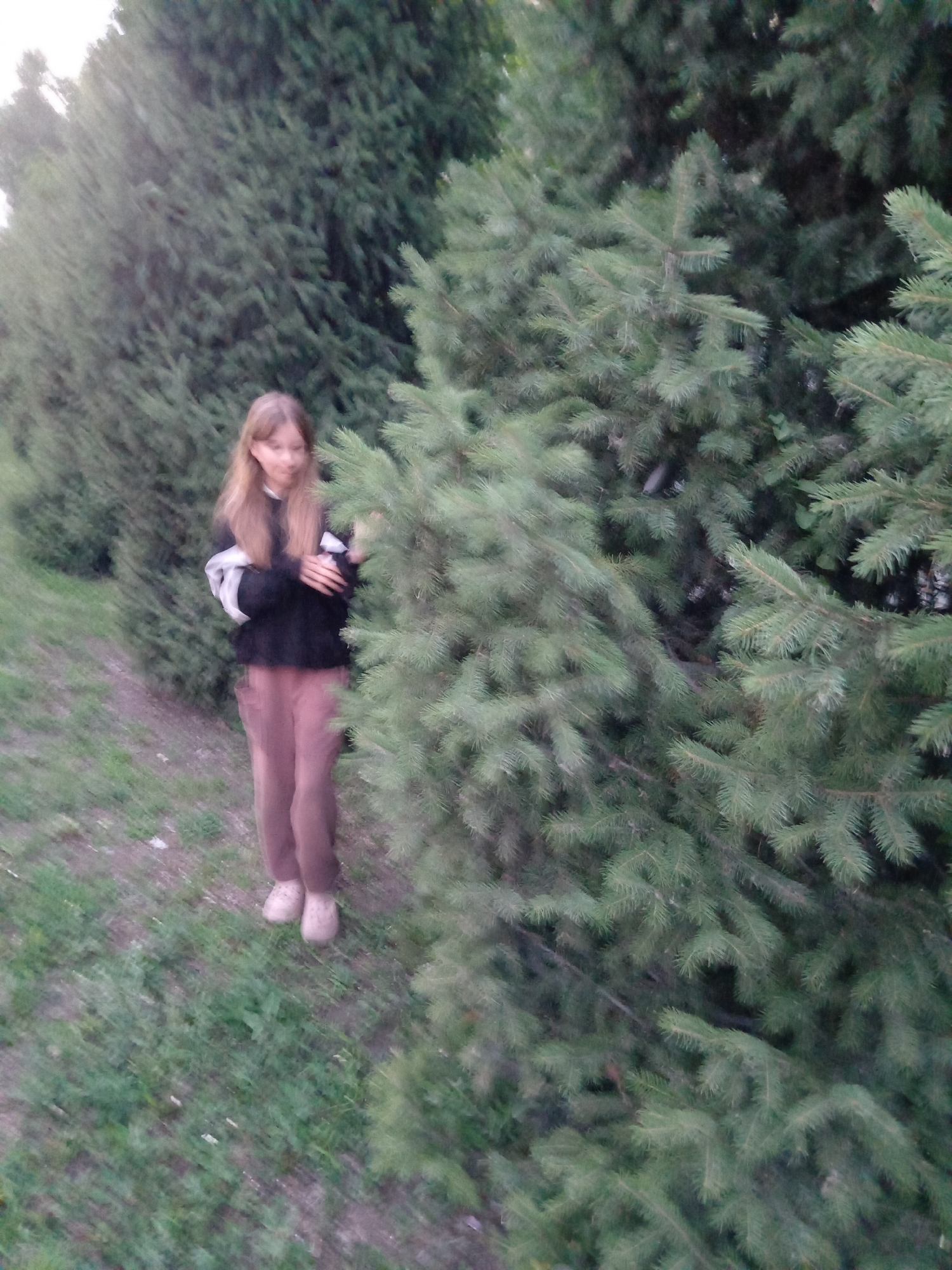 A young Caucasian girl with long blonde hair stands to the left of a large evergreen tree in a park setting. She is wearing a dark long-sleeved shirt with white stripes on the sleeves and light brown pants. Her arms are crossed in front of her. She is wearing light-colored shoes. The ground is covered in grass and some dirt. The background consists of more evergreen trees and foliage. The overall lighting suggests it is daytime, possibly late afternoon, with overcast skies. The image appears to be taken in Rudny, Kazakhstan, as suggested by the context.