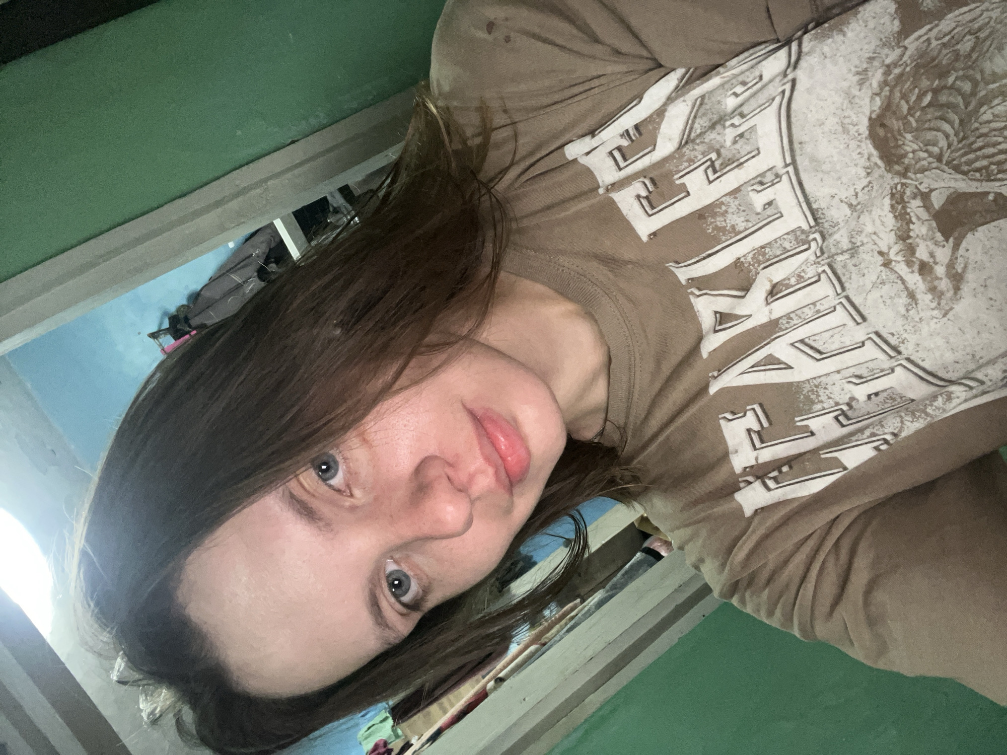 A young Caucasian female with medium brown hair and blue eyes is taking a selfie. She is wearing a tan t-shirt with a distressed graphic print. The background appears to be an interior with green and blue walls, and some hanging clothes are visible on the left. The scene is likely a domestic setting within the city of Nizhny Tagil, Russia.