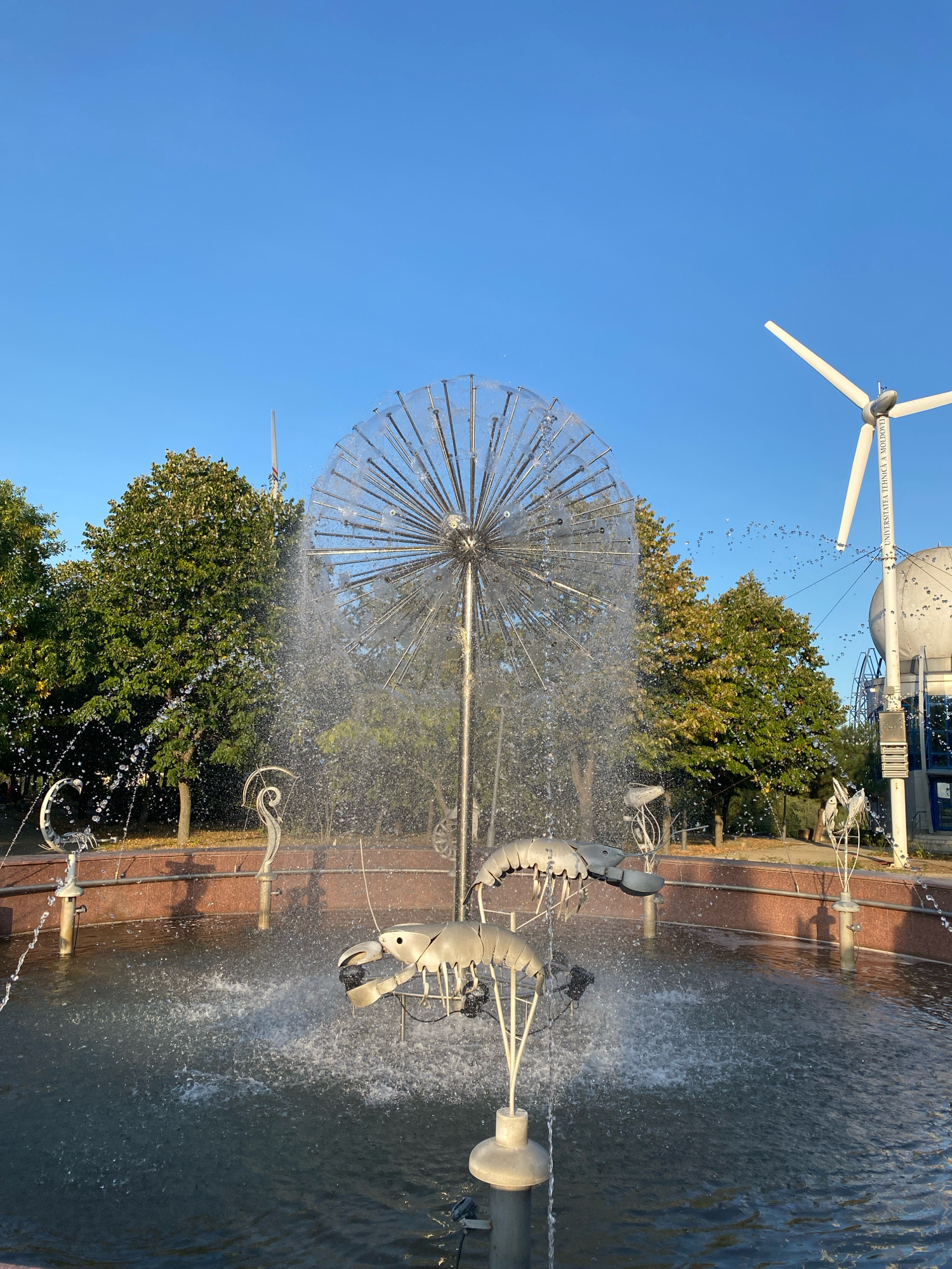 The image captures a fountain with kinetic sculptures of aquatic creatures in the foreground, set against a backdrop of lush green trees and a clear blue sky. To the right, a prominent white wind turbine stands tall, with a building partially visible beneath it. The wind turbine has text that reads "UNIVERSITATEA TEHNICĂ A MOLDOVEI". This suggests the location is on the grounds of the Technical University of Moldova in Chișinău, Moldova. The fountain itself features a large, spherical metal structure with numerous radiating spokes, from which water sprays, creating a misty effect. Several metal sculptures resembling shrimp or lobsters are positioned within the fountain's basin, with some appearing to interact with the water jets. The water is actively spraying from various points, creating a dynamic and lively scene. The surrounding area appears to be a park or campus setting, with trees and paved walkways visible. The sunlight suggests it is daytime, likely morning or afternoon, given the clear sky and shadows.