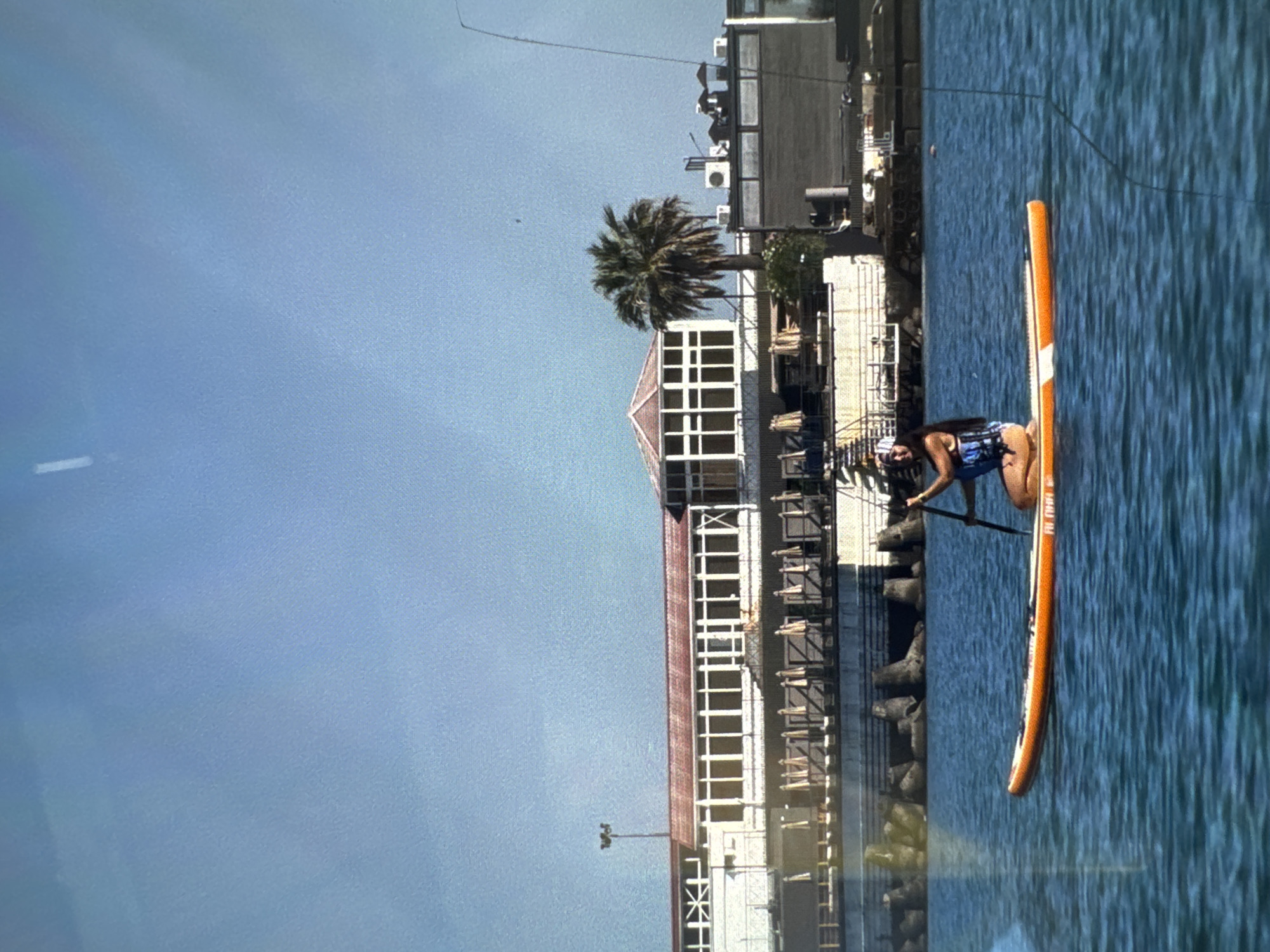 A woman is paddleboarding on a body of water. She is wearing a blue and white life vest and a patterned headscarf. She is kneeling on an orange and white paddleboard, holding a paddle. The water is a deep blue and appears calm. In the background, there is a building with a red roof, palm trees, and a clear blue sky. The building appears to be a resort or hotel, and the overall scene suggests a vacation or recreational setting in Sochi, Russia, possibly near the Black Sea.