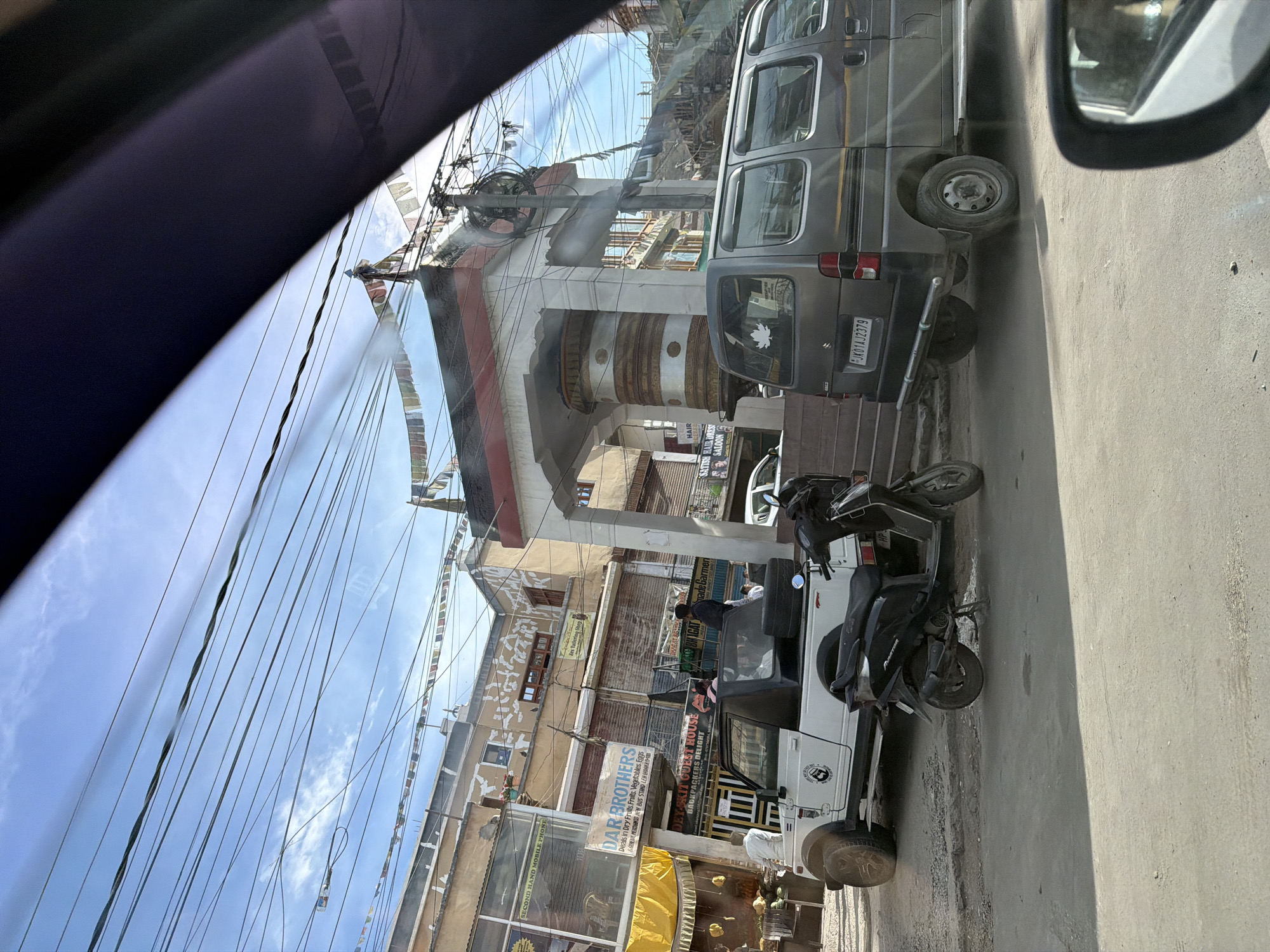 The image shows a street scene in the city of Leh, India, on a bright, clear day. In the foreground, a black scooter is parked on the side of the road. To its left, a white open-top jeep with people inside is also parked. Behind the jeep and scooter, a grey van is visible. Several buildings line the street, displaying various signs for businesses like "DAR BROTHERS" and "DEY-SKIT GUEST HOUSE". Prayer flags are strung across the street and on top of a large, cylindrical structure that appears to be a Buddhist prayer wheel. The sky is blue with scattered clouds, and numerous electrical wires crisscross overhead. The overall environment suggests a bustling urban setting within Leh.