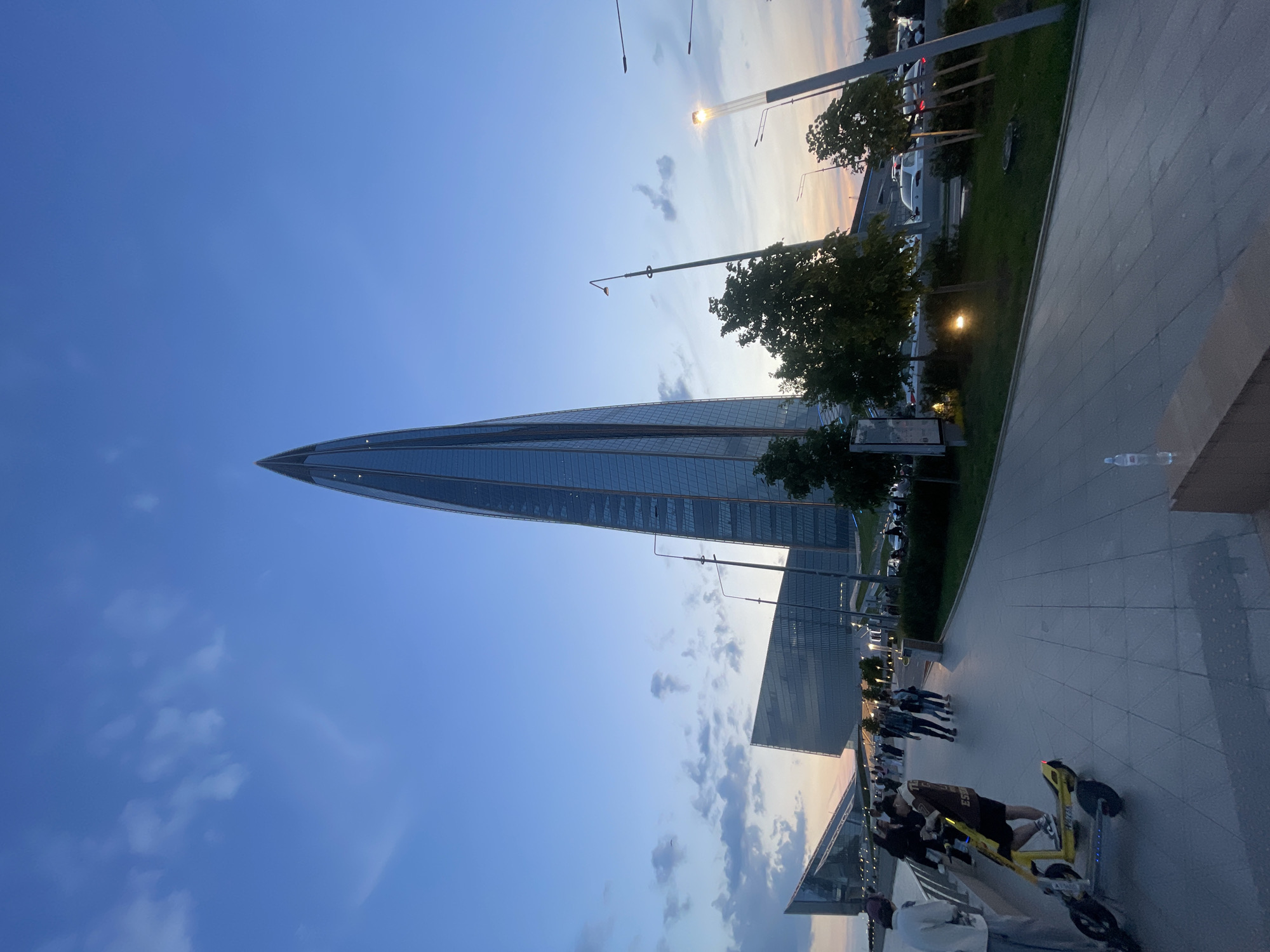 The image depicts the Lakhta Center in St. Petersburg, Russia, during twilight. A tall, spire-like skyscraper dominates the background, its glass facade reflecting the dusky sky. In the foreground, a wide paved walkway is occupied by several individuals, some walking and others standing. A yellow electric scooter is parked on the left side of the walkway. Adjacent to the walkway is a grassy area with scattered trees and streetlights. The sky is a gradient of blue and orange, with wispy clouds. Cars are visible on a road in the middle ground.