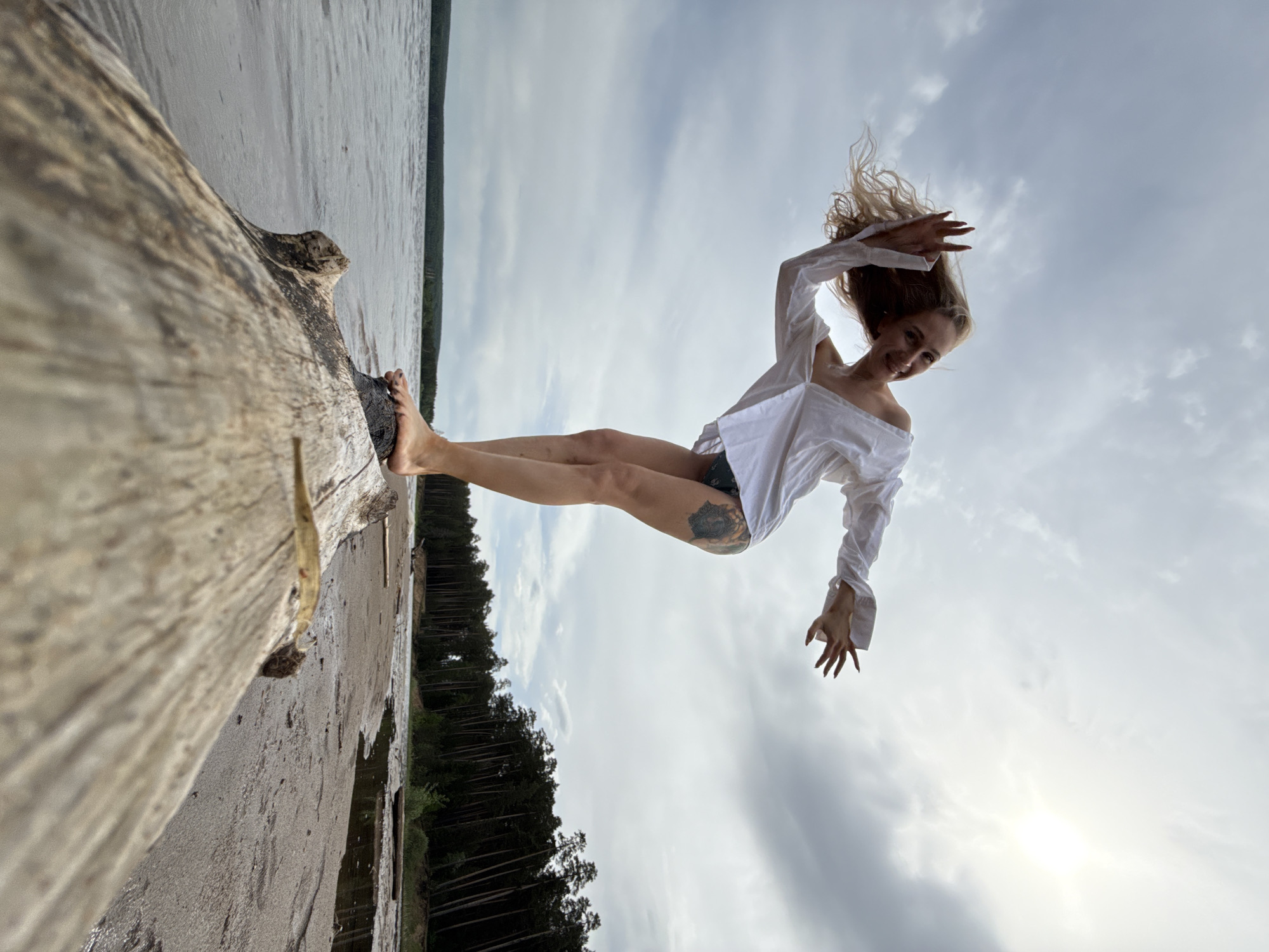 A woman with fair skin and blonde hair is positioned horizontally in the frame, appearing to levitate against a cloudy sky. She is wearing a white, off-the-shoulder blouse and dark shorts, and a tattoo is visible on her upper thigh. Her body is angled with her feet resting on a fallen log or tree trunk that dominates the left side of the image. The background includes a body of water and a forest line. The location is likely the city of Chaikovsky, Russia.