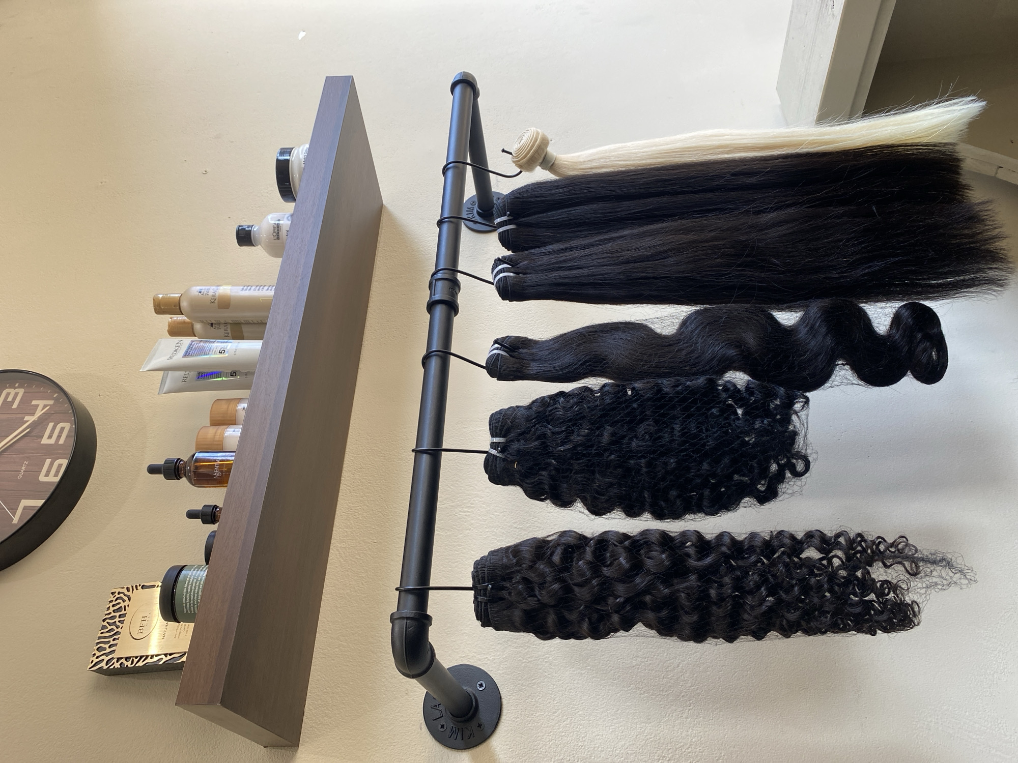 This media file captures an indoor setting, likely a professional hair salon or retail space, displaying hair extensions and hair care products against a light-colored wall.
The primary subjects are various bundles of hair extensions and a collection of hair care products. On the left side of the image, a dark brown wooden shelf is mounted on the wall, holding several bottles of hair care products. These products include brands such as "L'OREAL PROFESSIONNEL," "KERACARE," and "REDKEN," with some bottles featuring dropper tops. A product from "BFH NATURAL" is also visible on the bottom shelf. A portion of a round black wall clock, labeled "QUARTZ," can be seen on the far left.
To the right of the shelf, a black industrial-style pipe rack is vertically mounted on the wall. Hanging from this rack are five distinct bundles of hair extensions, secured by thin black hooks. From top to bottom, these bundles include a long, straight, platinum blonde extension; a long, straight, black extension; a bundle of wavy or loose curly black hair; a bundle of tight curly or kinky black hair partially enclosed in a black mesh net; and a final bundle of long, spiral curly black hair also secured in a black mesh net. The pipe fitting at the bottom of the rack has some text stamped on it, partially visible as "KIM WIX."
The scene depicts a well-organized display of hair products and extensions, suggesting an activity related to hair styling, sales, or professional use. The lighting is bright and even, indicating either ample natural light or strong artificial illumination, characteristic of a professional environment. No people are visible, and there are no discernible location cues to suggest a specific city or country.