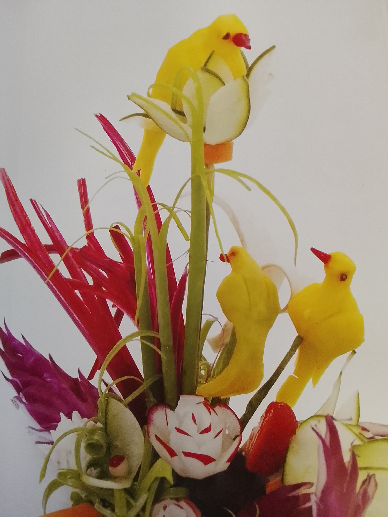 The image displays a vegetable arrangement featuring sculpted yellow birds, likely crafted from yellow peppers or squash. The birds have red pepper beaks and are perched on stems of what appear to be scallions or other similar vegetables. The arrangement also incorporates red and purple cabbage leaves, carved radishes resembling roses, and zucchini slices. The scene is set against a plain white background. The location in Altagracia, Venezuela is unknown.