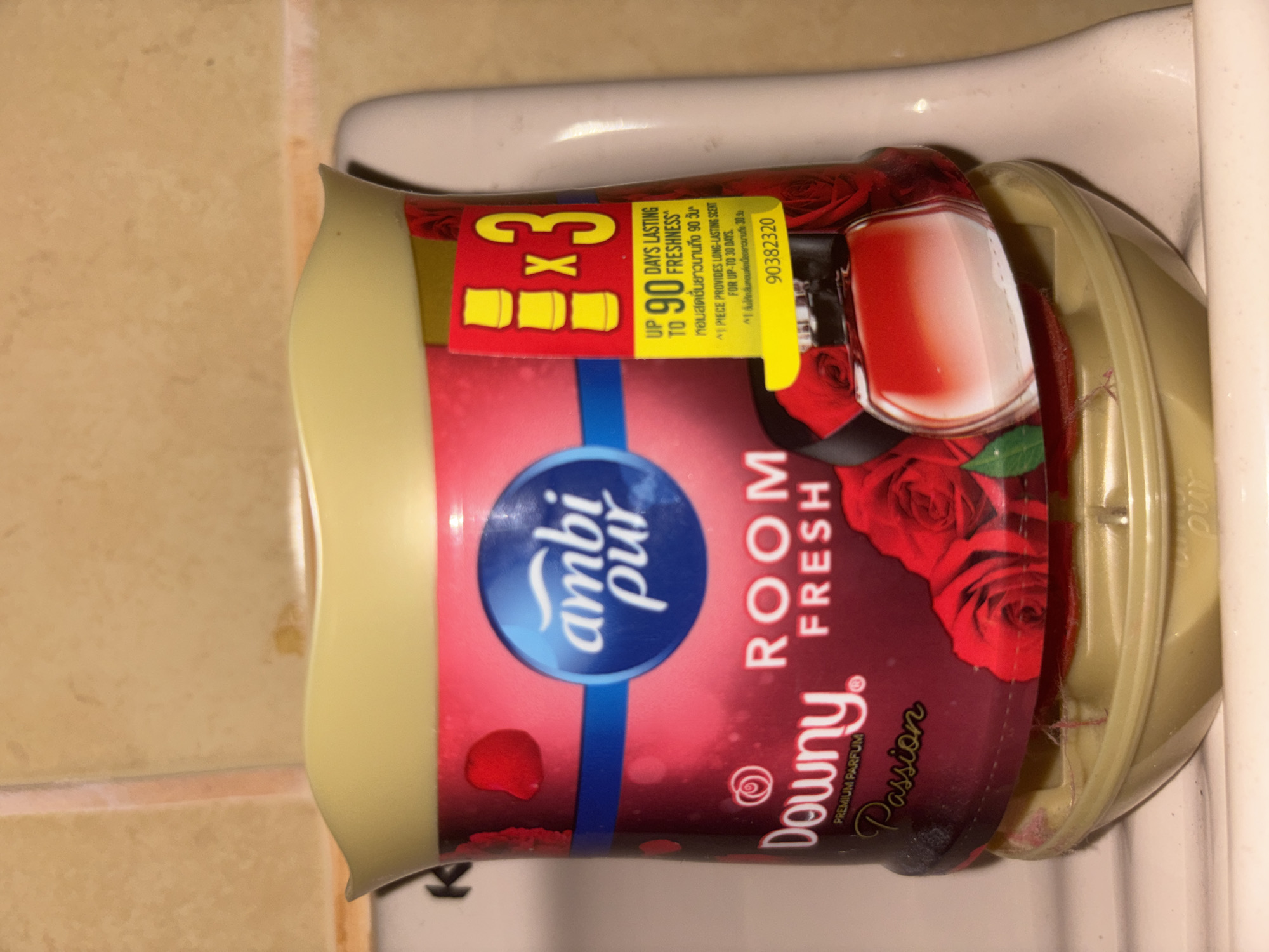 The image shows a product labeled "Downy Room Fresh Passion" placed on a light-colored ceramic surface, likely a sink or toilet tank. The product is a red and gold container with a yellow lid. The label features the brand names "Ambi Pur" and "Downy," along with images of roses and a perfume bottle. Text on the label indicates "Up to 90 days lasting freshness" and includes a product code. The background consists of tiled walls. The lighting suggests an indoor environment. There are no people or actions depicted. The location is identified as Chalong, Thailand.