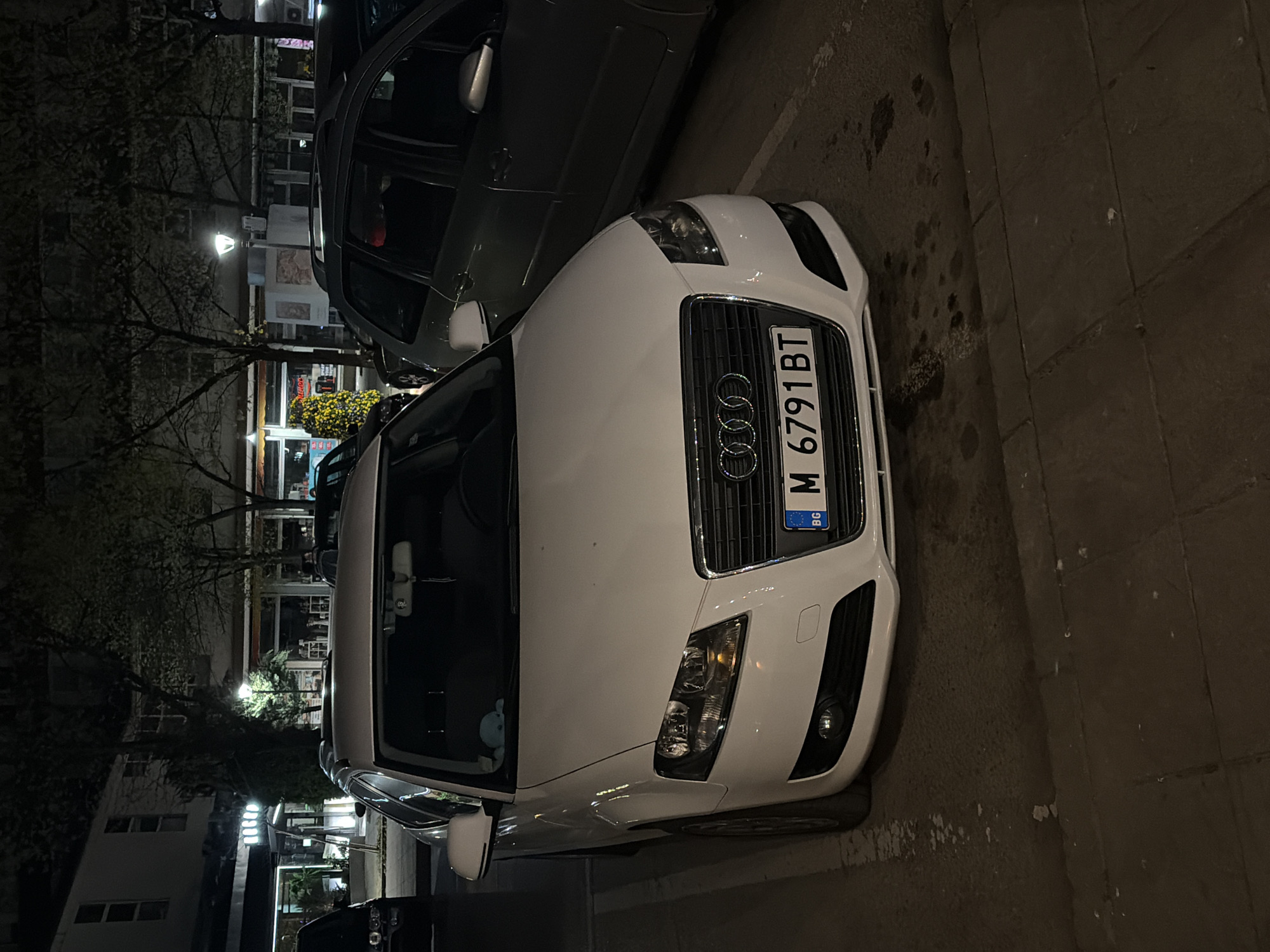 The image shows a white Audi car parked on a street in Montana, Bulgaria. The car's license plate is "M 6791 BT". The vehicle is parked on the right side of the street, which is paved with stone. A gray SUV is parked alongside. The background includes buildings, trees, streetlights, and some storefronts, suggesting an urban or town center. The image was likely taken at night or in low-light conditions.