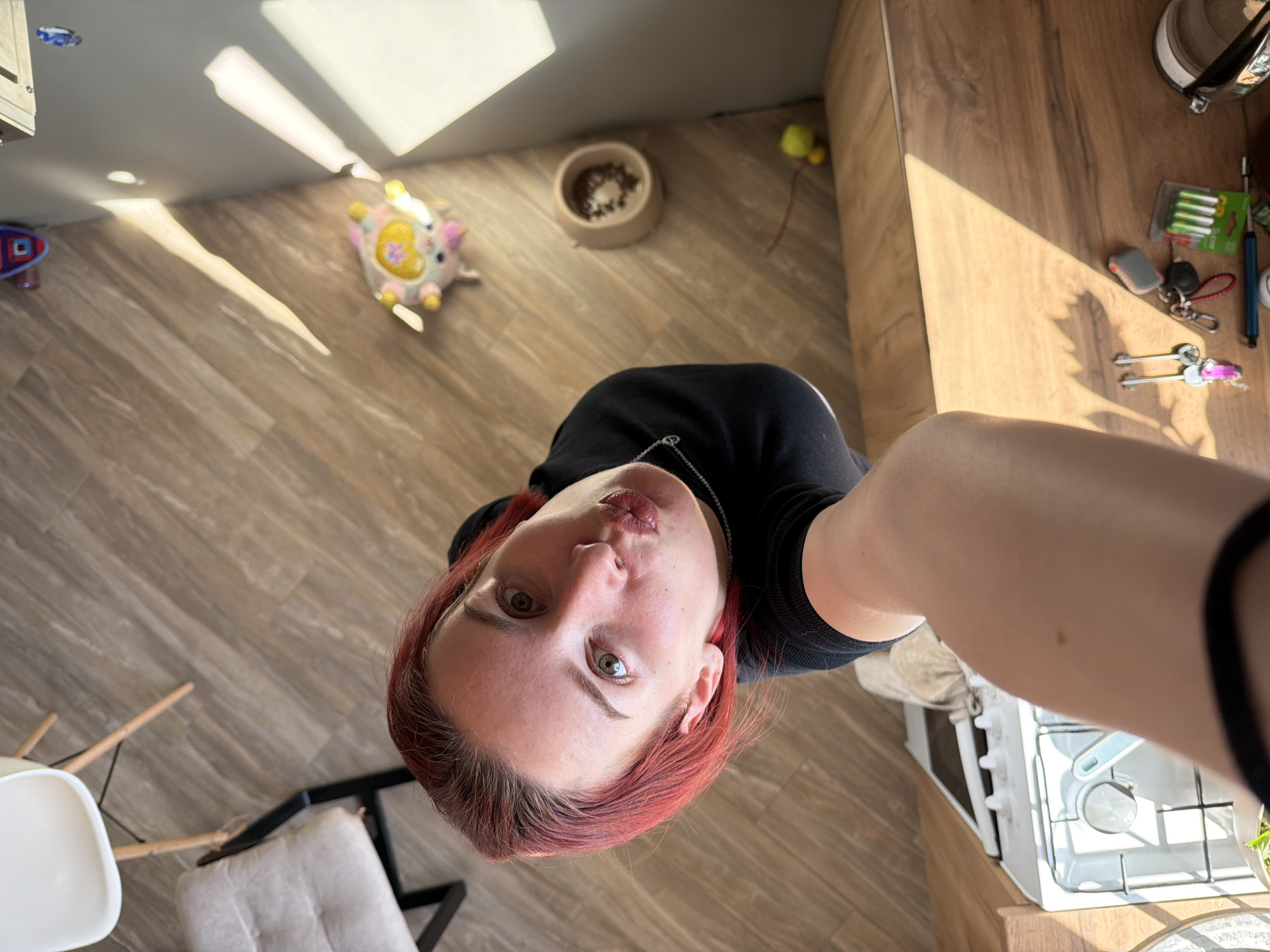 A high-angle, close-up selfie shows a person with pale skin, red hair, and green eyes. The person wears a black top and a silver chain necklace. The background reveals a domestic interior with a brown wood-patterned floor. A child's toy, a pet food bowl, and various items on a countertop are visible, including keys, batteries, and cooking utensils near a stove. The location is identified as Chaikovsky, Russia, based on available metadata.