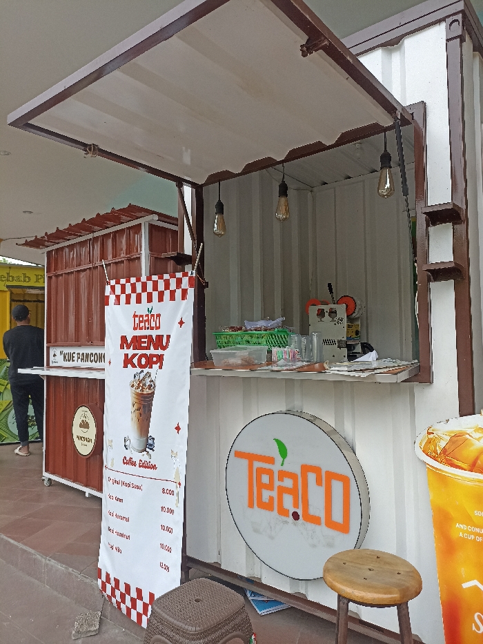 This daytime image, taken in Tulungagung, Indonesia, captures an outdoor food and drink stall setup, likely in a market or commercial area.
The primary subject is a white container-style stall for a business named "TeaCo," with its front window propped open to serve customers. Inside, a cup sealer machine, plastic cups, straws, and other beverage preparation items are visible on the counter. Edison-style light bulbs hang from the ceiling. A circular backlit sign bearing the "TeaCo" logo with a green leaf is mounted on the front. To its right, a large graphic of an iced drink, also with the "TeaCo" logo, shows partially visible text like "AND CONU..." and "A CUP OF...". Two stools, one brown and one darker plastic, are placed in front of the stall.
A tall white banner, adorned with a red and white checkered pattern at the top and bottom, hangs from the "TeaCo" stall. It displays the "teaco MENU KOPI" (Coffee Menu) under "Coffee Edition." The menu lists:
- Original Kopi Susu (Original Milk Coffee) - 8.000
- Kopi Aren (Palm Sugar Coffee) - 10.000
- Kopi Karamel (Caramel Coffee) - 10.000
- Kopi Hazelnut (Hazelnut Coffee) - 10.000
- Kopi Milo (Milo Coffee) - 12.000
(Prices are likely in Indonesian Rupiah.)
To the left of the "TeaCo" stall is a smaller brown stall, also appearing to be a vendor booth, with a sign that reads "KUE PANCONG" (a type of Indonesian cake). A man, dressed in dark clothing, stands with his back to the camera near this brown stall. The overall setting is brightly lit, suggesting clear weather conditions.