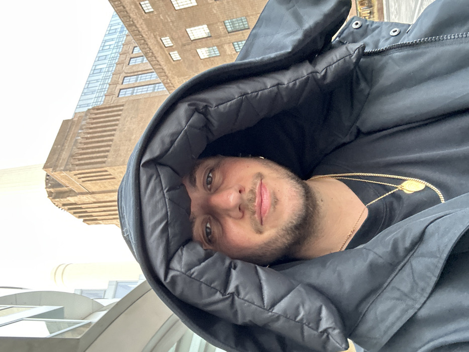A person with light skin, a short beard, and a black hooded jacket looks at the camera. He's wearing two gold chain necklaces and gold earrings. The photo is taken outdoors with buildings in the background. The composition is angled, giving a perspective of looking up at the subject. The sky appears overcast.