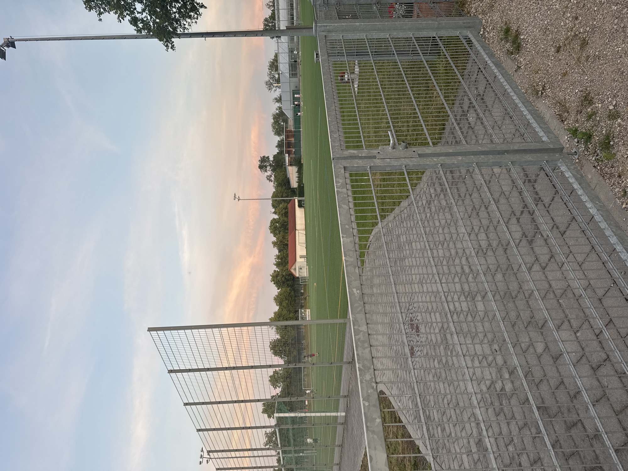 An outdoor, low-angle, full shot captures a view of a football pitch in Neubiberg, Germany, during what appears to be sunset or twilight. The sky is a soft blend of pale blue and pastel pinks, with scattered clouds. In the foreground, a galvanized metal fence with a gate separates a paved walkway from a grassy area. The walkway is made of grey interlocking pavers. Beyond the fence, the green football pitch is visible, marked with yellow lines. Several figures, appearing to be young people, are scattered across the field, some engaged in activities that suggest a practice or casual game. In the background, there are trees, a small building with a red roof, and stadium lighting poles. The overall scene depicts a sports facility at the end of a day.