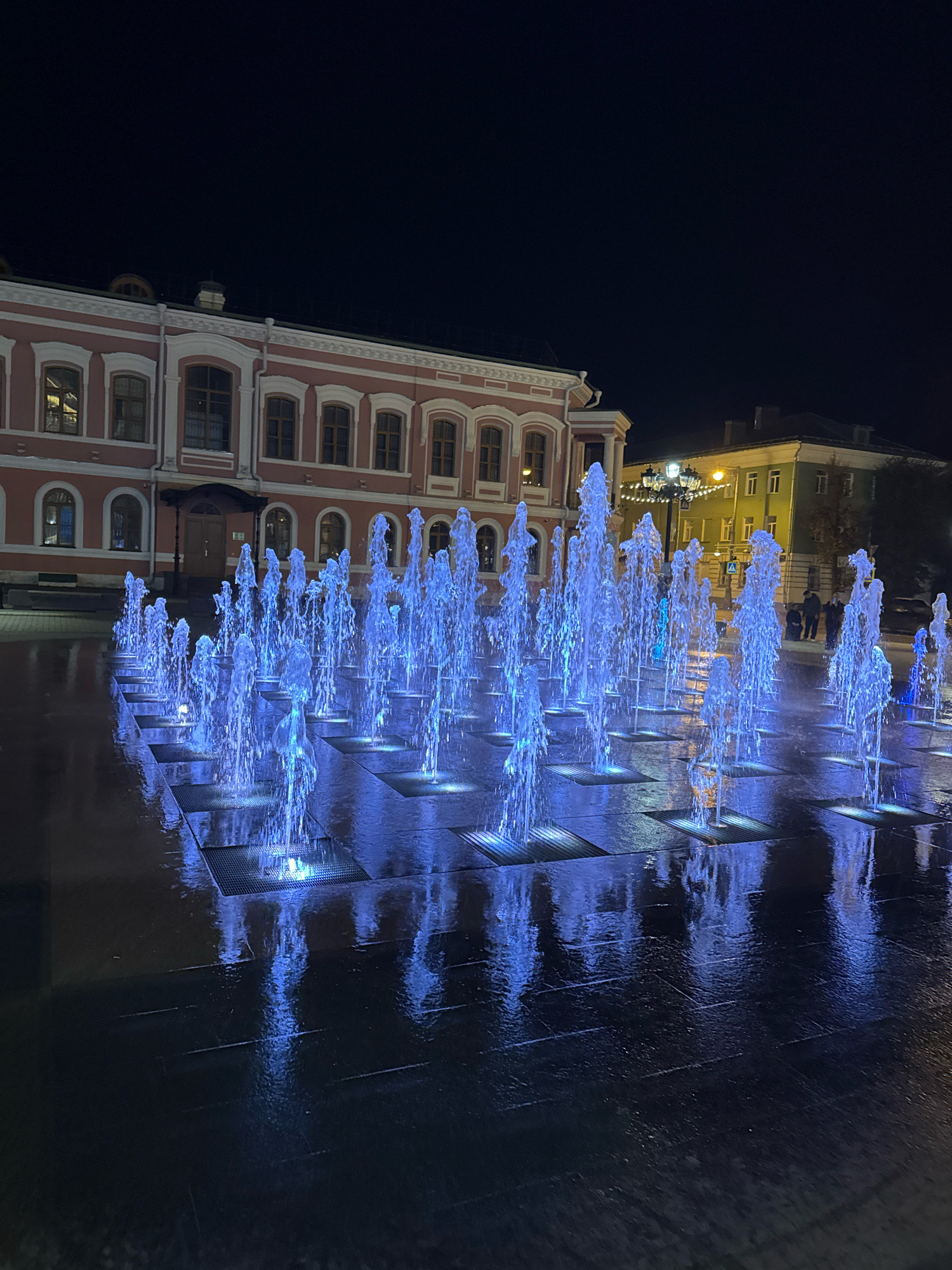 This is a nighttime photograph of a modern fountain installation in Tver, Russia. The fountain features numerous jets of water illuminated with blue light, creating a vibrant display against the dark sky and wet pavement. In the background, several buildings are visible, including a large, historic pink building with arched windows and ornate trim on the left, and a smaller green building with lit windows on the right. Streetlights and some decorative lights are also present in the background. A few people are seen standing in the distance near the green building. The ground around the fountain is wet, reflecting the blue light from the water and the surrounding lights, suggesting it may have recently rained or the fountain has been recently operated. The overall scene depicts an urban public space illuminated at night.