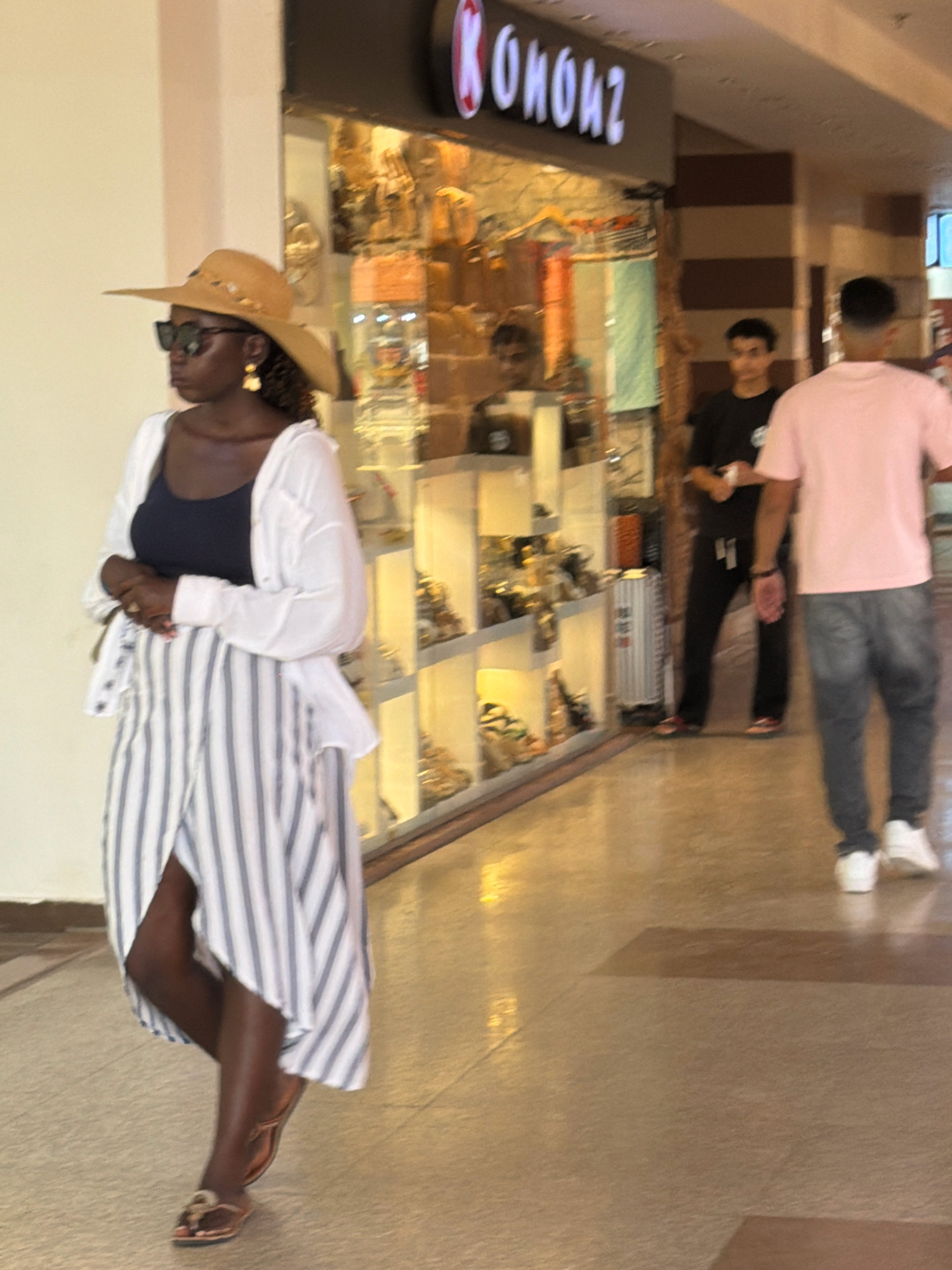 A woman wearing a wide-brimmed hat, sunglasses, a dark tank top, a white open-buttoned shirt, and a striped skirt walks past a storefront in Sharm El Sheikh, Egypt. The storefront displays various items behind glass shelves. Two men are visible in the background, one wearing a black t-shirt and dark pants, the other a pink t-shirt and grey jeans. The environment appears to be an indoor shopping area with a tiled floor.