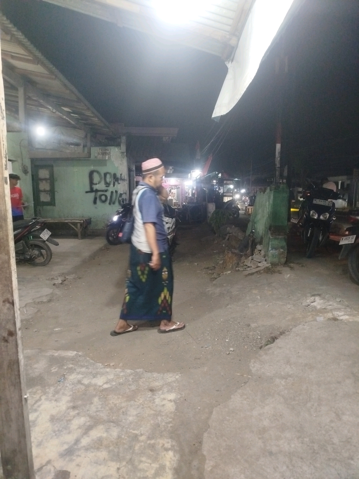 The image depicts a nighttime scene on a street in Burneh, Indonesia. A man wearing a pink peci (a traditional cap), a blue shirt, and a sarong is walking away from the camera. He has a bag slung over his shoulder. In the background, there are several motorcycles parked. One motorcycle in the foreground on the left is dark colored with a red accent. Another motorcycle, dark colored with a license plate that reads "W 6233 HJ", is prominent in the middle ground. Further in the background, more motorcycles are visible, some with their headlights on. The street is paved with asphalt and gravel. There are some buildings with green walls and wooden roofs. Graffiti reading "DON TONG" is visible on one of the walls. The environment appears to be a commercial or residential area with open stalls or shops. The lighting is dim, suggesting artificial illumination from streetlights and shop signs.