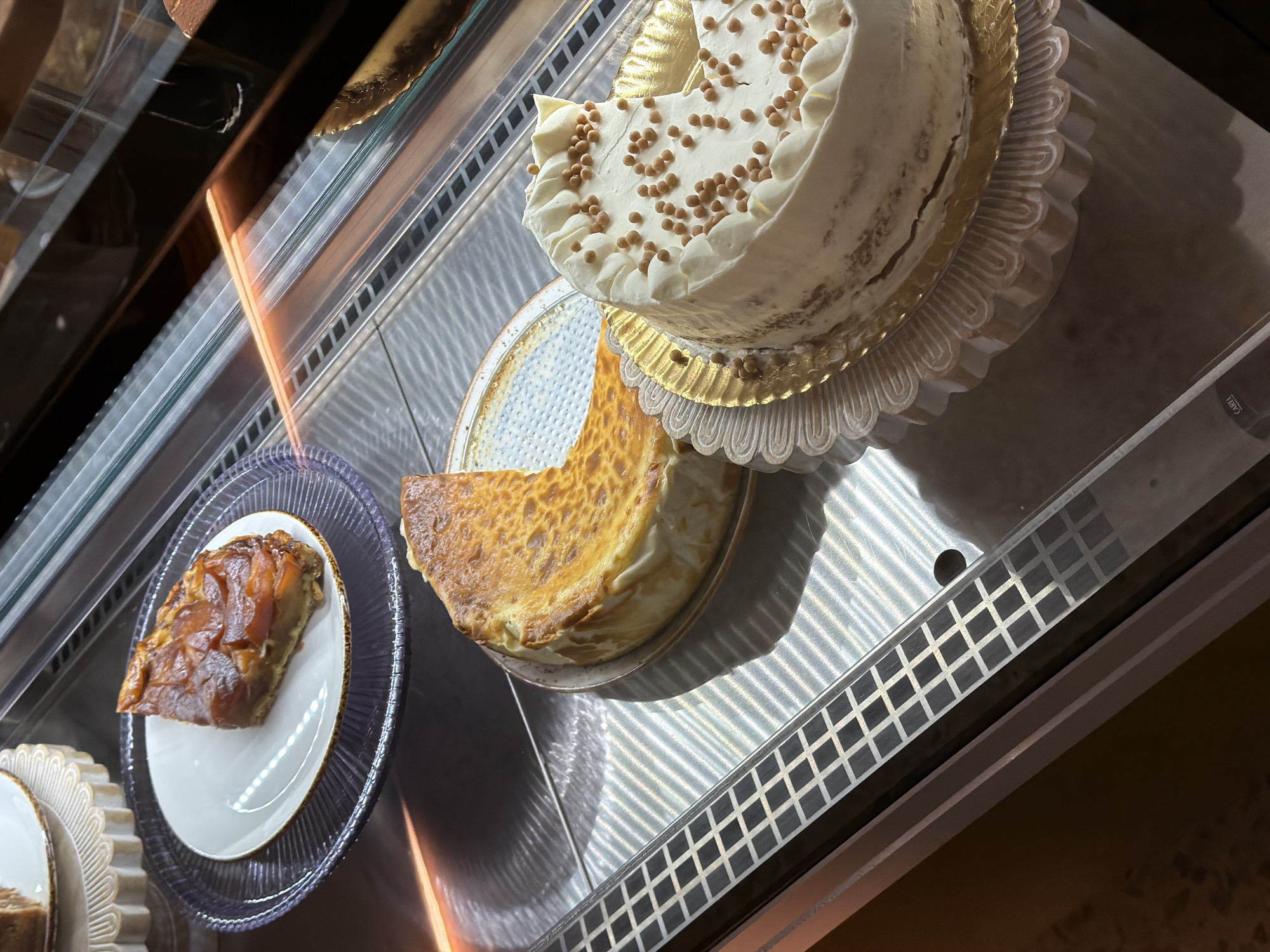 The image displays a close-up view of a dessert display case, likely in a bakery or cafe in Benidorm, Spain. In the foreground, a slice of white frosted cake with small, caramel-colored spheres is visible. To the left of this cake, a half-eaten cheesecake sits on a plate. Further left, a slice of apple tart is presented on a white plate, which is itself on a fluted glass stand. The display case features a metallic ribbed surface and glass panels, with internal lighting casting reflections and shadows on the desserts. No people are present in the immediate scene.
