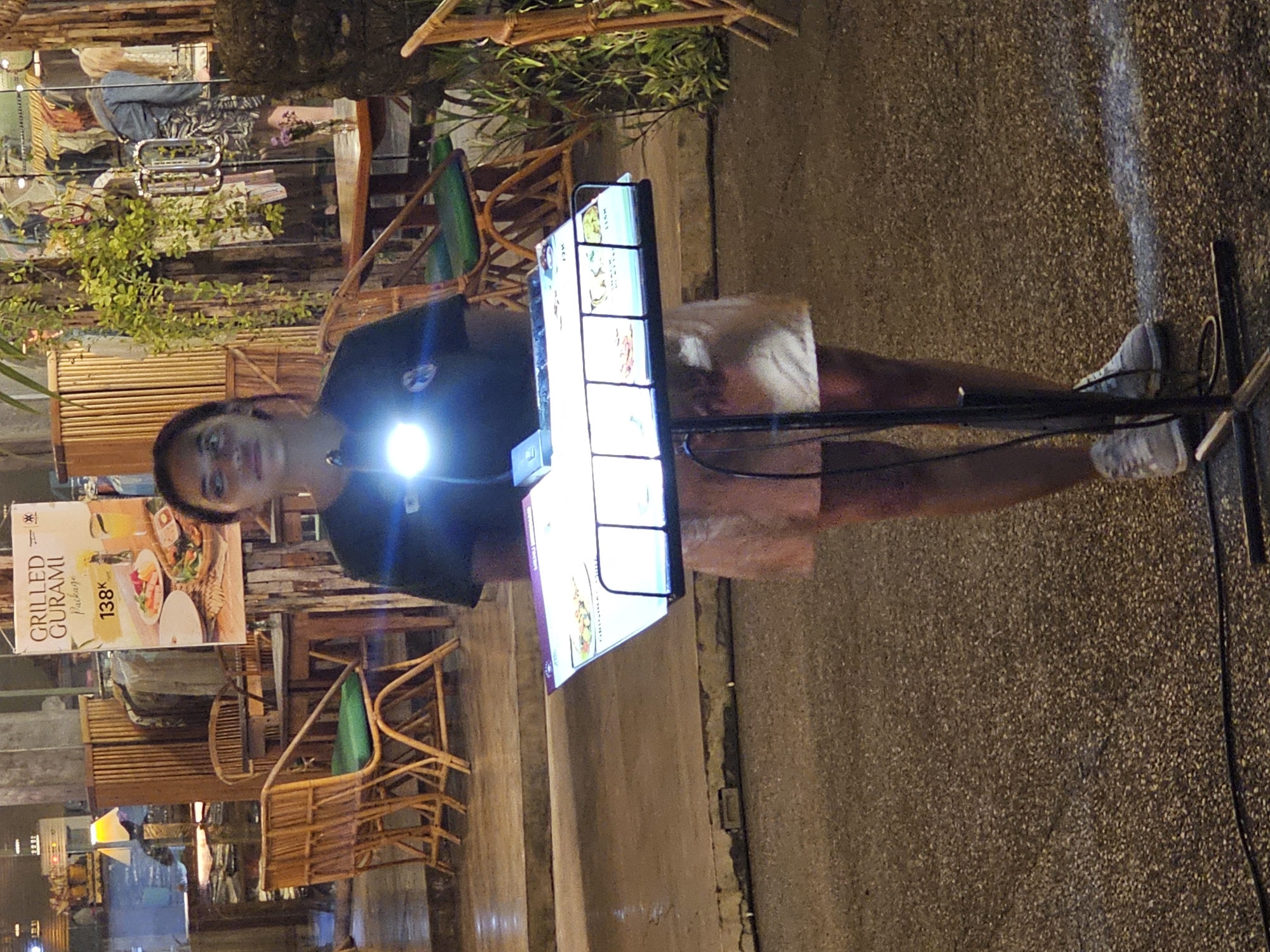 The image captures a street scene in Denpasar, Indonesia. A person stands on the right, facing the camera, wearing a dark t-shirt and a light-colored skirt or shorts, with athletic shoes. In front of the person is a multi-panel display menu, likely for a restaurant or food establishment. The menu is illuminated and showcases various food items.
The setting includes outdoor seating with wooden chairs and tables on the left. A sign advertising "Grilled Gurami" for 138K is visible in the background, further suggesting a restaurant setting. The ground is paved, and a power cord is seen running along the ground near the person's feet. The scene is illuminated by artificial lighting, creating a warm ambiance. The overall impression is a vibrant outdoor dining area, common in urban environments like Denpasar.