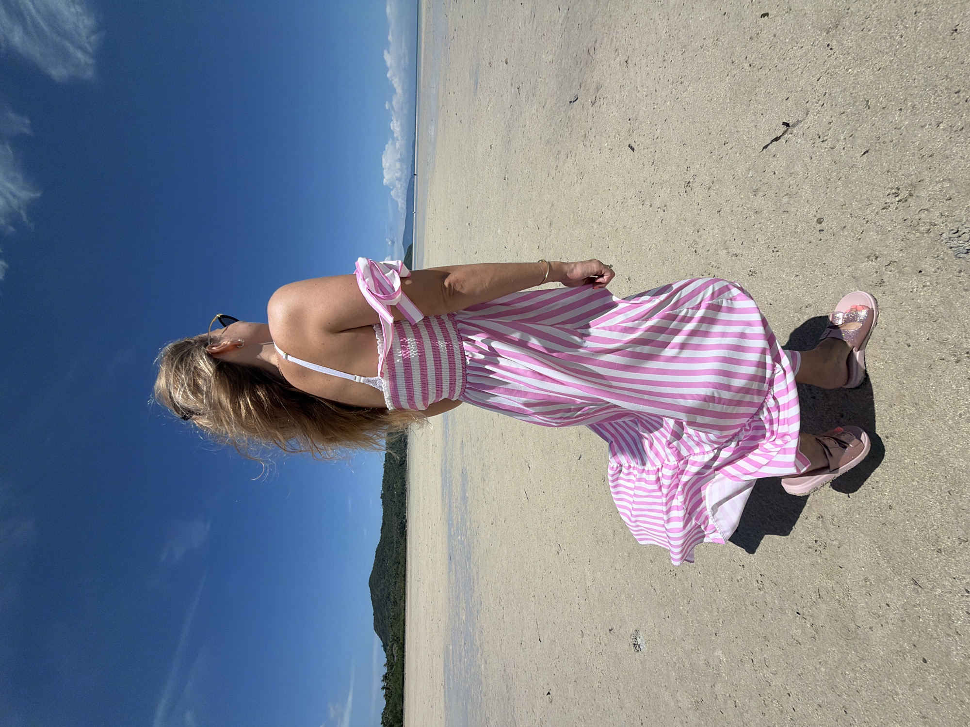 In ตำบลเกาะพะงัน, Thailand, a person stands on a sandy beach under a clear blue sky with scattered clouds. The individual is facing away from the camera, looking out towards the ocean. They are wearing a pink and white striped dress with off-the-shoulder sleeves and pink sandals. The dress is flowing in the wind. The beach is wide and appears to be low tide, with shallow water visible in the distance. A green, tree-covered island or peninsula is visible on the horizon. The lighting suggests it is daytime, likely mid-morning or afternoon.