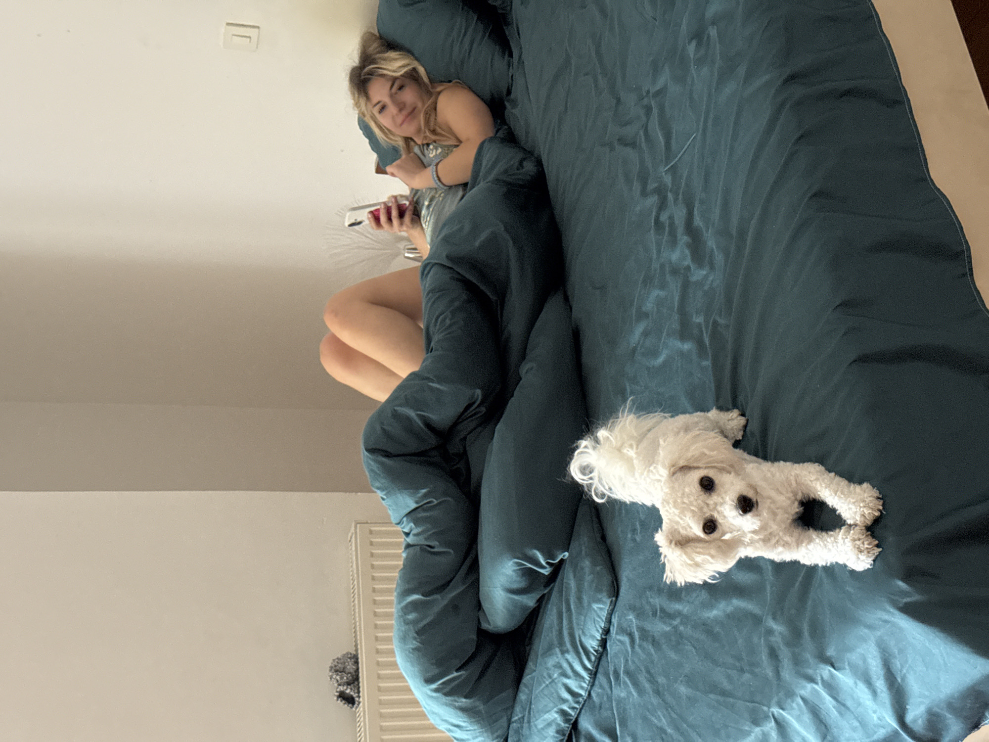 A woman is lying in a bed covered with a blue duvet, holding a mobile phone. A small white dog is lying on the bed next to her. The location appears to be an interior room, possibly in Budva, Montenegro, though this is not definitively determinable from the image. A radiator is visible on the wall. A light switch is also visible on the ceiling.