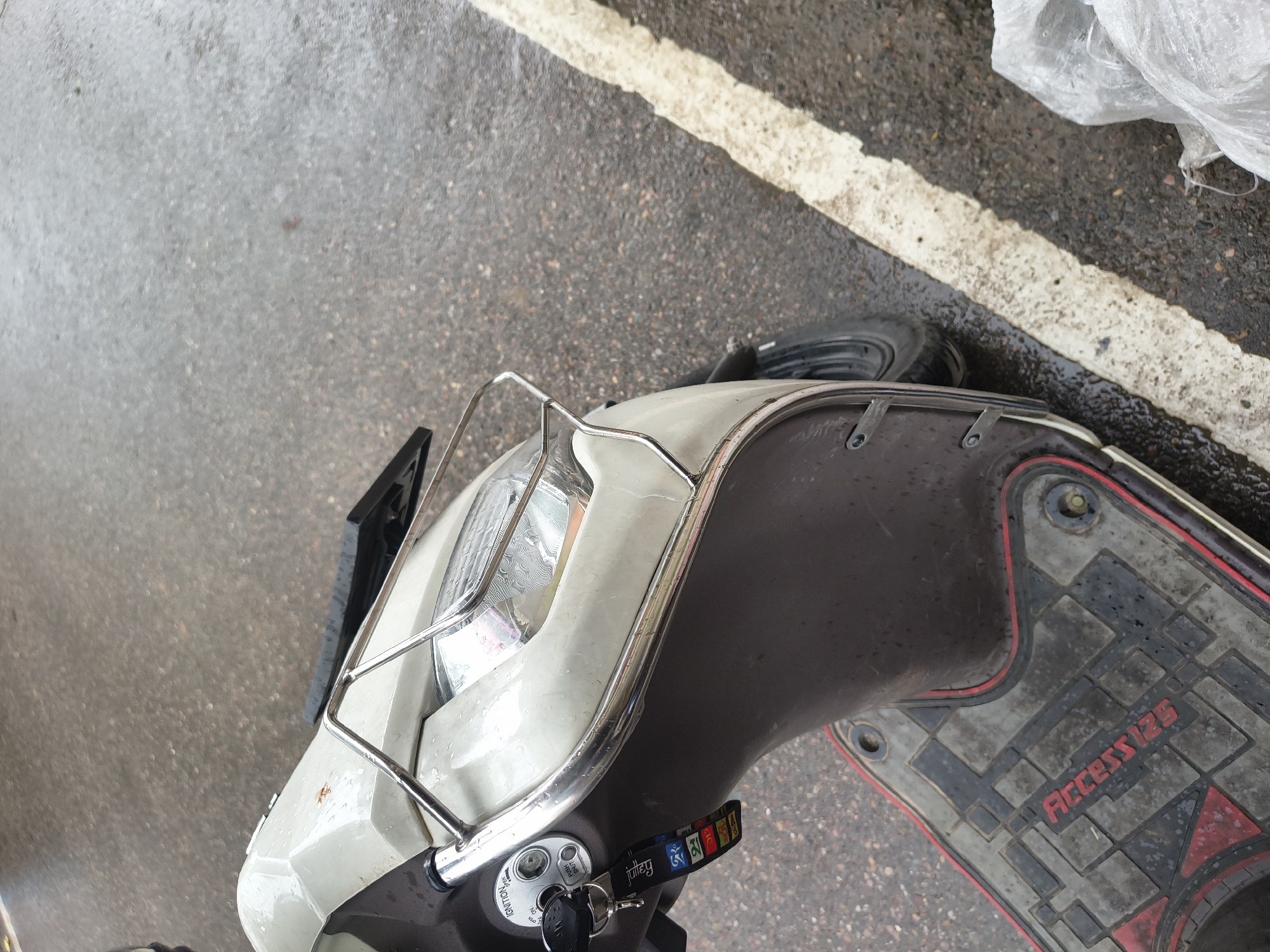 A close-up, slightly angled, medium shot captures the front portion of a light-colored scooter parked on a wet, grey asphalt road. The scooter has a chrome guard over its front, and a black ignition switch with a key and a colorful fabric keychain are visible. The footboard of the scooter, colored grey and black with red trim, has the text "Access 125" embossed in red. A white painted line runs along the right side of the road. To the far right, a portion of a translucent white plastic bag is visible. The overall scene suggests an outdoor environment in Dharamshala, India, possibly during or after rainfall, indicated by the wet road surface.