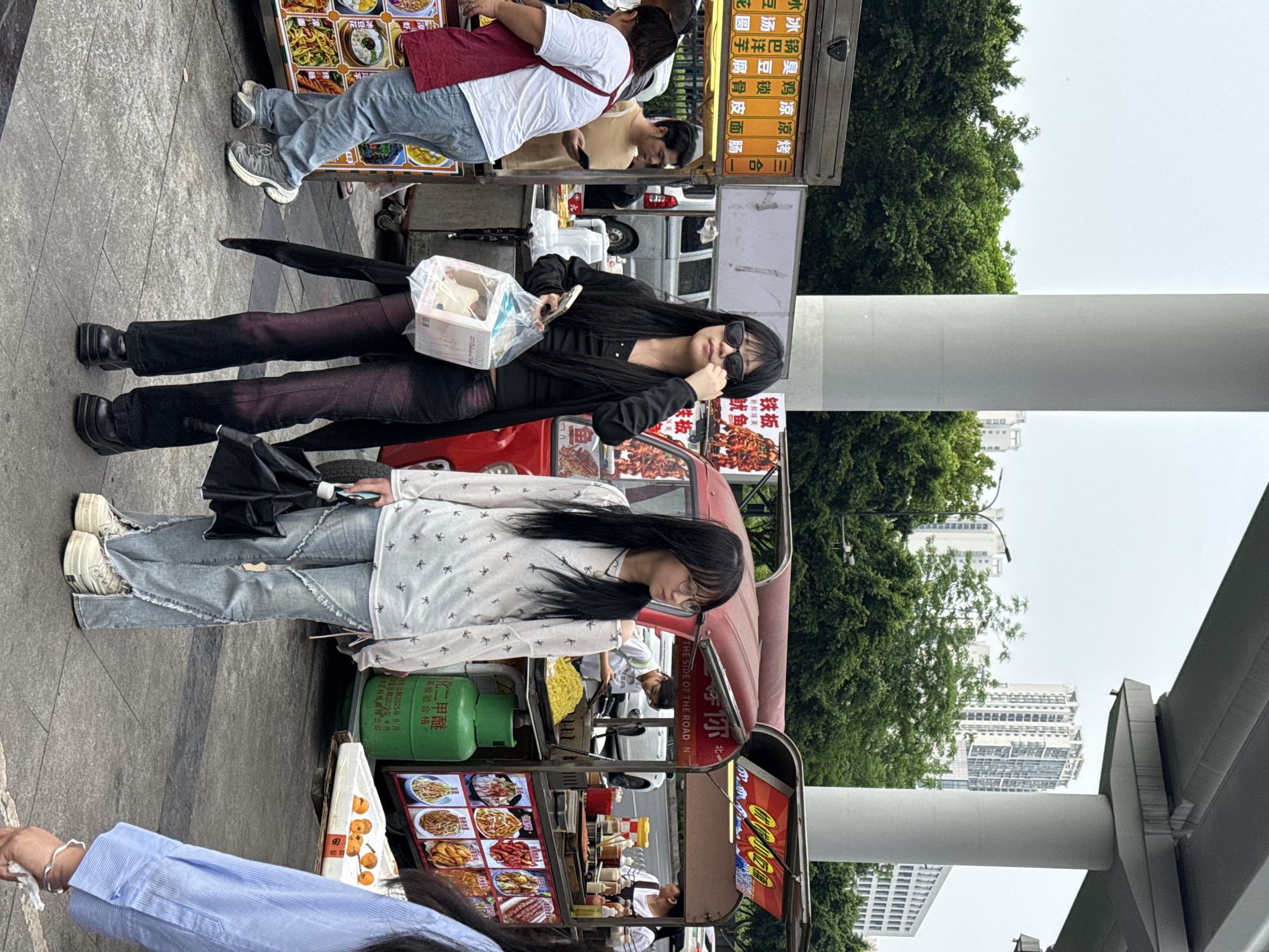 The image depicts a street scene in the Chongqing High-tech Zone, China. Two young women stand next to street food vendors. One woman, dressed in a black coat, is holding a plastic container and adjusting her sunglasses. Another young woman wears a patterned white blouse and jeans. Behind her is a green propane tank and a street food stall with various food items on display. A second food stall is visible behind the first young woman, with a menu in Chinese characters. Buildings are visible in the background.