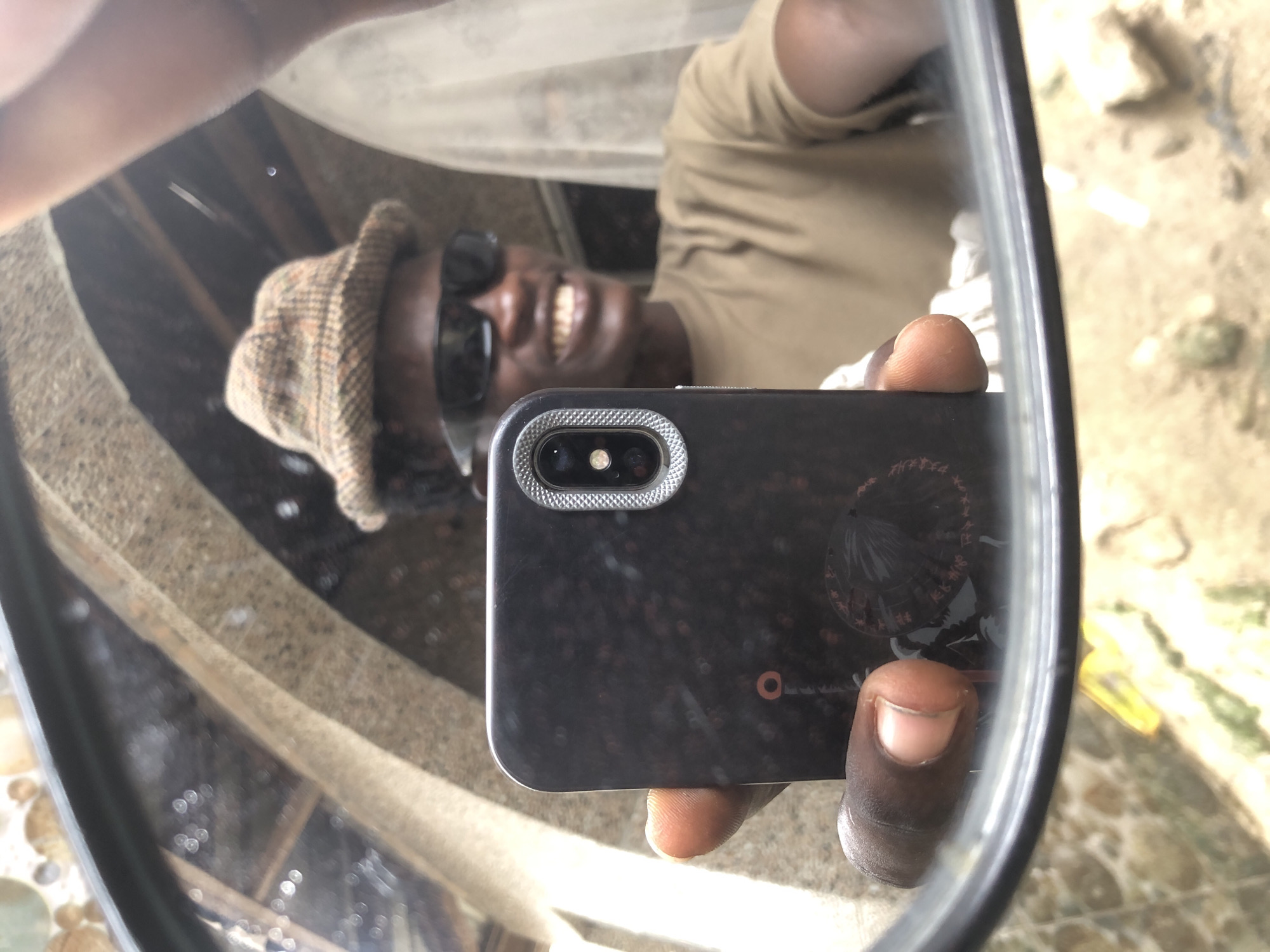 A young Black male is taking a mirror selfie, holding a smartphone in his left hand to capture his reflection. He is wearing a brown plaid bucket hat and black sunglasses, and a light brown t-shirt. The background suggests an outdoor setting, possibly in Ajah, Nigeria, with textured walls and an arched structure visible. The reflection shows his smiling face and the camera lens of his phone. The mirror's frame and the surrounding environment are also visible in the reflection.