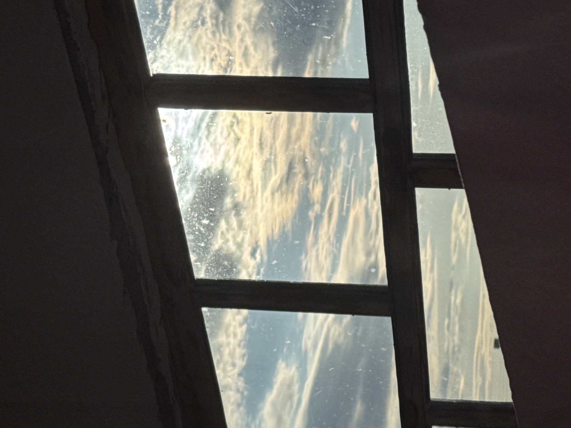 The media file presents an interior view looking out through a multi-paned window, likely situated in Chaikovsky, Russia. The window features a dark, possibly wooden, frame that divides the scene into several rectangular glass sections. The window panes appear dirty, streaked, or dusty, with visible imperfections and possibly water marks.
Through the window, a cloudy sky is visible. The clouds are illuminated by bright, diffused light, displaying a range of white, golden, and light blue hues. The warm tones and the way the light catches the clouds suggest either an early morning sunrise or a late afternoon sunset. The light highlights the textures of the clouds, creating a dynamic appearance.
The immediate surroundings inside the room, on the left and right of the window frame, are dark and indistinct, likely showing parts of a wall or window casing. There are no people visible, nor any specific activities taking place. No text is discernible within the image. The scene conveys a sense of quiet observation of the sky from an indoor perspective.