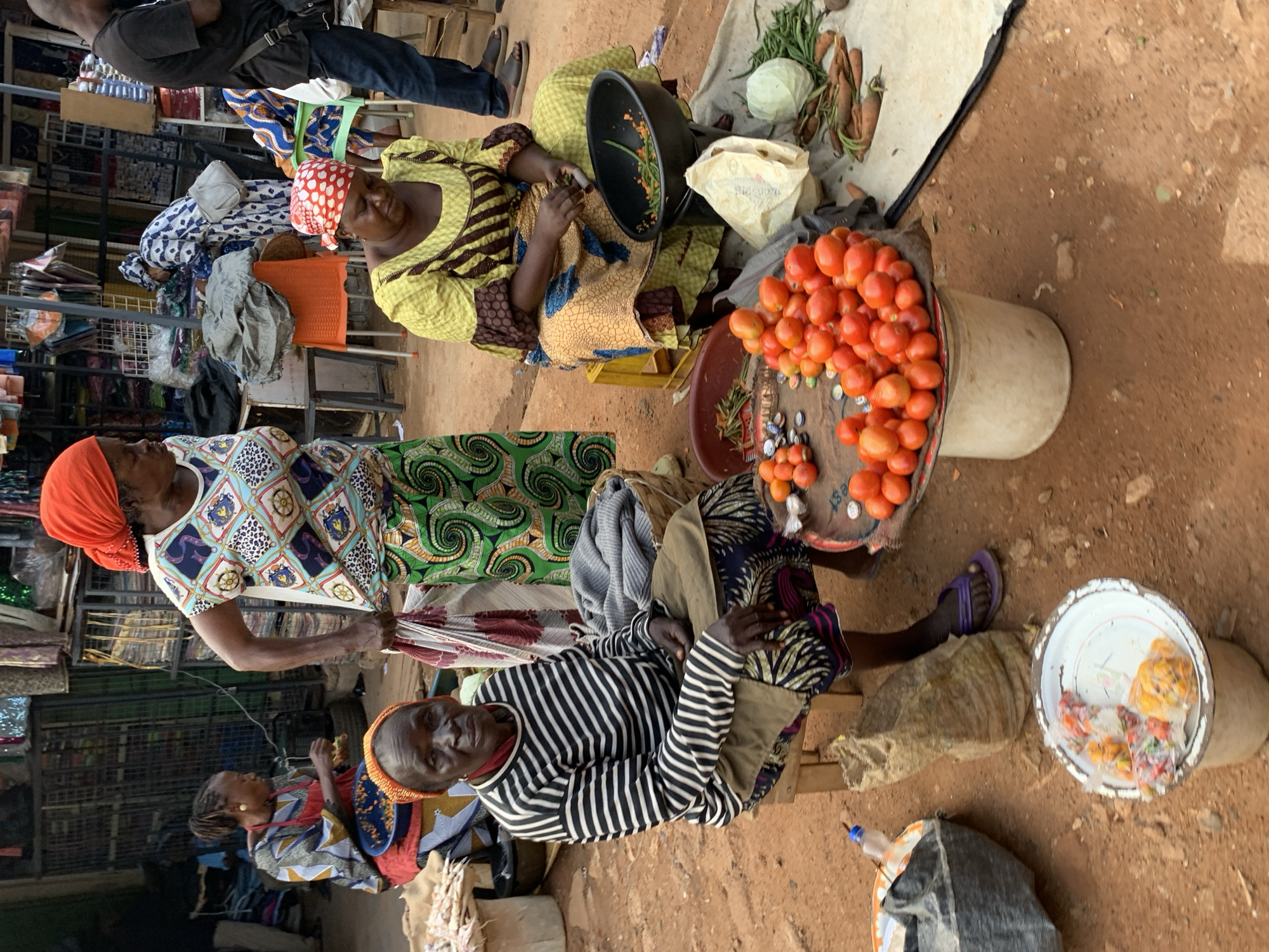 In Jos, Nigeria, a market scene unfolds as several individuals, primarily women, engage in commerce. The subjects are predominantly women adorned in vibrant, patterned clothing and headscarves. One woman in a yellow and brown striped dress, wearing a red and white polka dot headscarf, sits beside a black bowl containing chopped vegetables and a partially visible white paper bag. Another woman in a colorful patterned dress and an orange headscarf stands, her hands occupied with fabric. A third woman, dressed in a black and white striped shirt and a red headscarf, is seated on a wooden stool, her gaze directed towards the viewer.
Objects of note include a large bucket overflowing with ripe red tomatoes, a shallow bowl with what appear to be packaged food items, and a variety of vegetables such as carrots and green beans displayed on a cloth. Various fabrics and materials are visible in the background, suggesting a bustling marketplace with diverse goods.
The scene appears to be taking place during daylight hours, with natural light illuminating the market stalls and the ground, which is made of reddish-brown earth. The atmosphere seems to be one of active trade and daily life in a Nigerian market. The expressions on the faces of the women suggest a sense of resilience and engagement in their work. No visible text is discernible in the image.