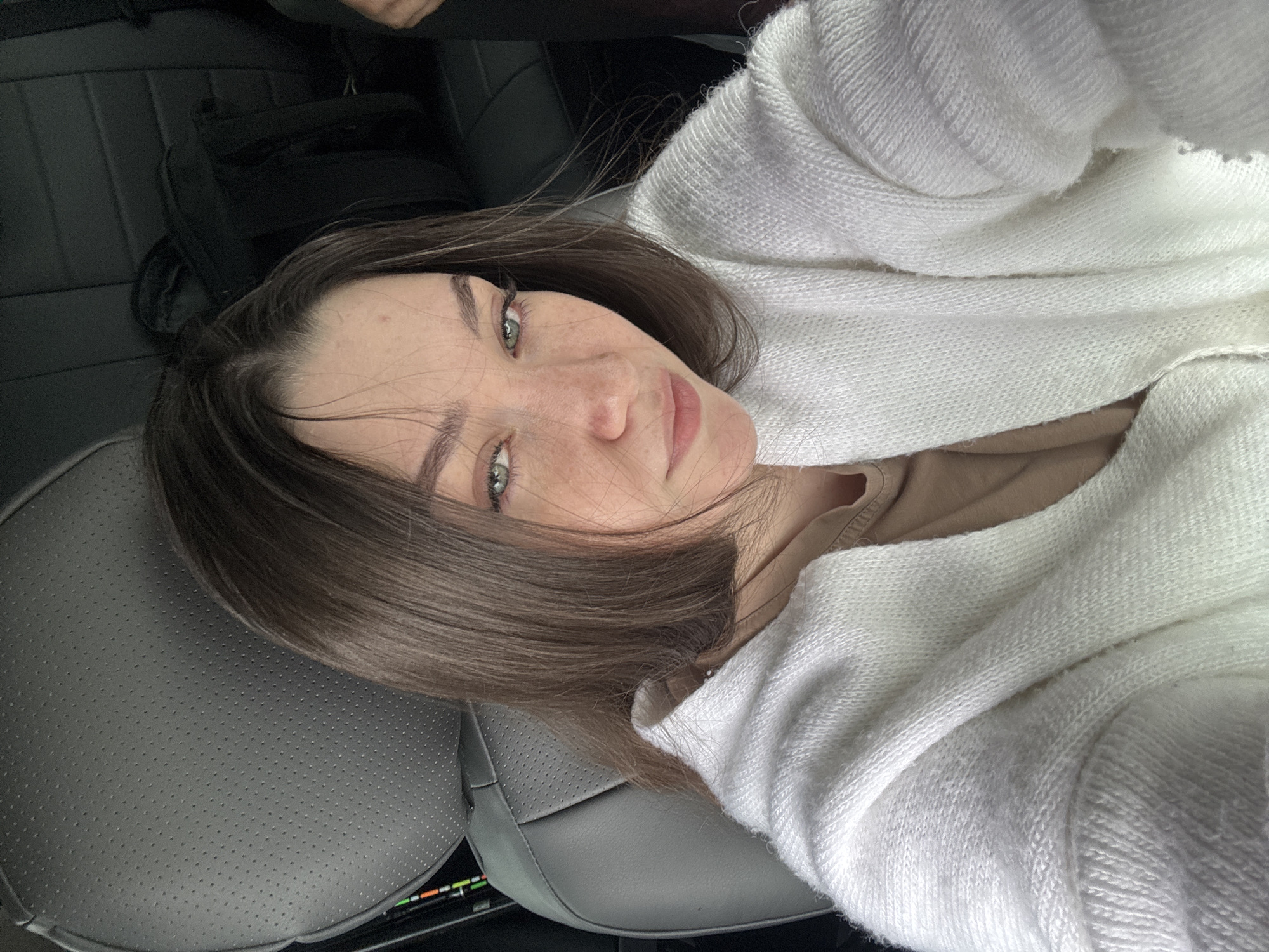 The image shows a young woman with shoulder-length brown hair and green eyes, wearing a white knitted sweater over a tan top. She is sitting in the passenger seat of a car, evidenced by the grey perforated leather headrest and the dark interior of the vehicle. Her face is turned towards the camera, and she has a neutral expression with a slight tilt of her head. She has freckles across her nose and cheeks. The background shows the dark interior of a car, with a seat and some bags visible. No specific location in Perm, Russia can be identified from the visual information provided.