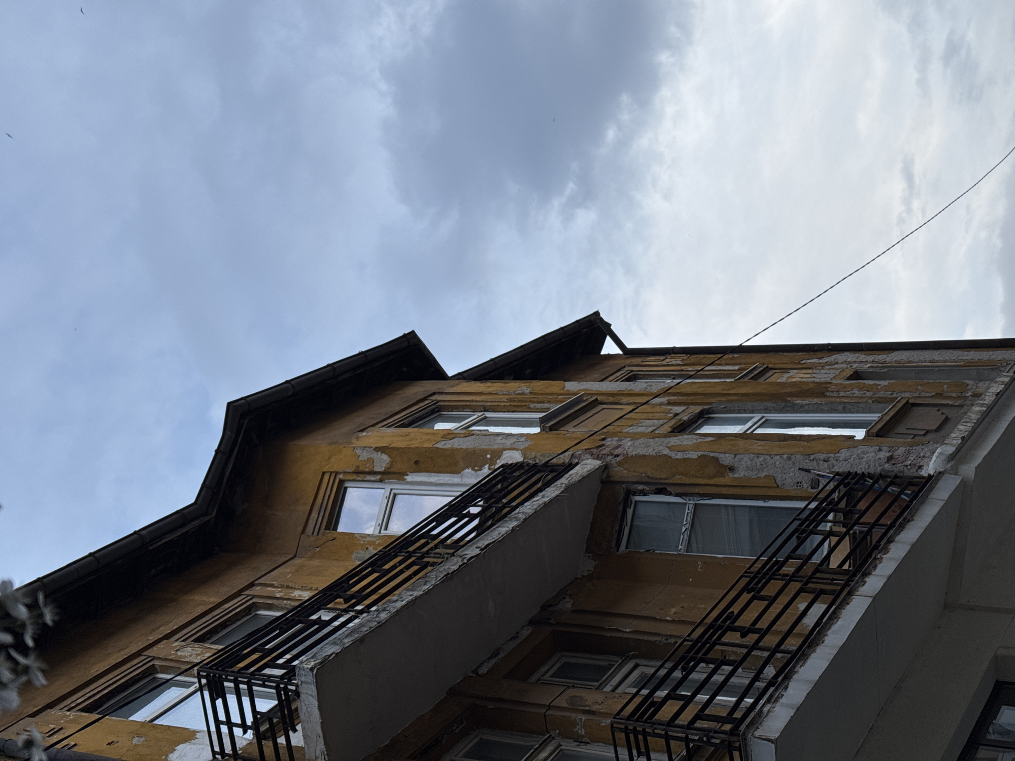 The image offers an upward-looking perspective of an old, multi-story building facade set against an overcast, cloudy sky. Located in Sofia, Bulgaria, the building's exterior is painted in a prominent yellow or ochre hue, which is extensively weathered and peeling, exposing lighter plaster or brickwork beneath.
Numerous rectangular windows with white or light-colored frames are integrated into the facade, reflecting the dull, grey sky. Several dark, gridded metal structures, appearing to be rudimentary balconies or window safety bars, protrude from the building's face, contributing to its aged aesthetic. A dark roofline with distinct eaves is visible along the top edges of the building, indicating an upper level or attic.
A thin, dark utility wire or cable stretches across the upper right section of the sky. Small, indistinct dark specks, possibly distant birds, can be faintly observed against the light sky in some sections. Blurry, light-colored foliage partially frames the bottom left corner of the main image, suggesting nearby vegetation.
The scene captures a static moment in an urban environment during daytime, characterized by cloudy weather and a sense of historical wear. There is no discernible human activity, nor is any text visible on the building or within the frame. The overall impression is one of an aged structure in need of maintenance, common in older city districts.