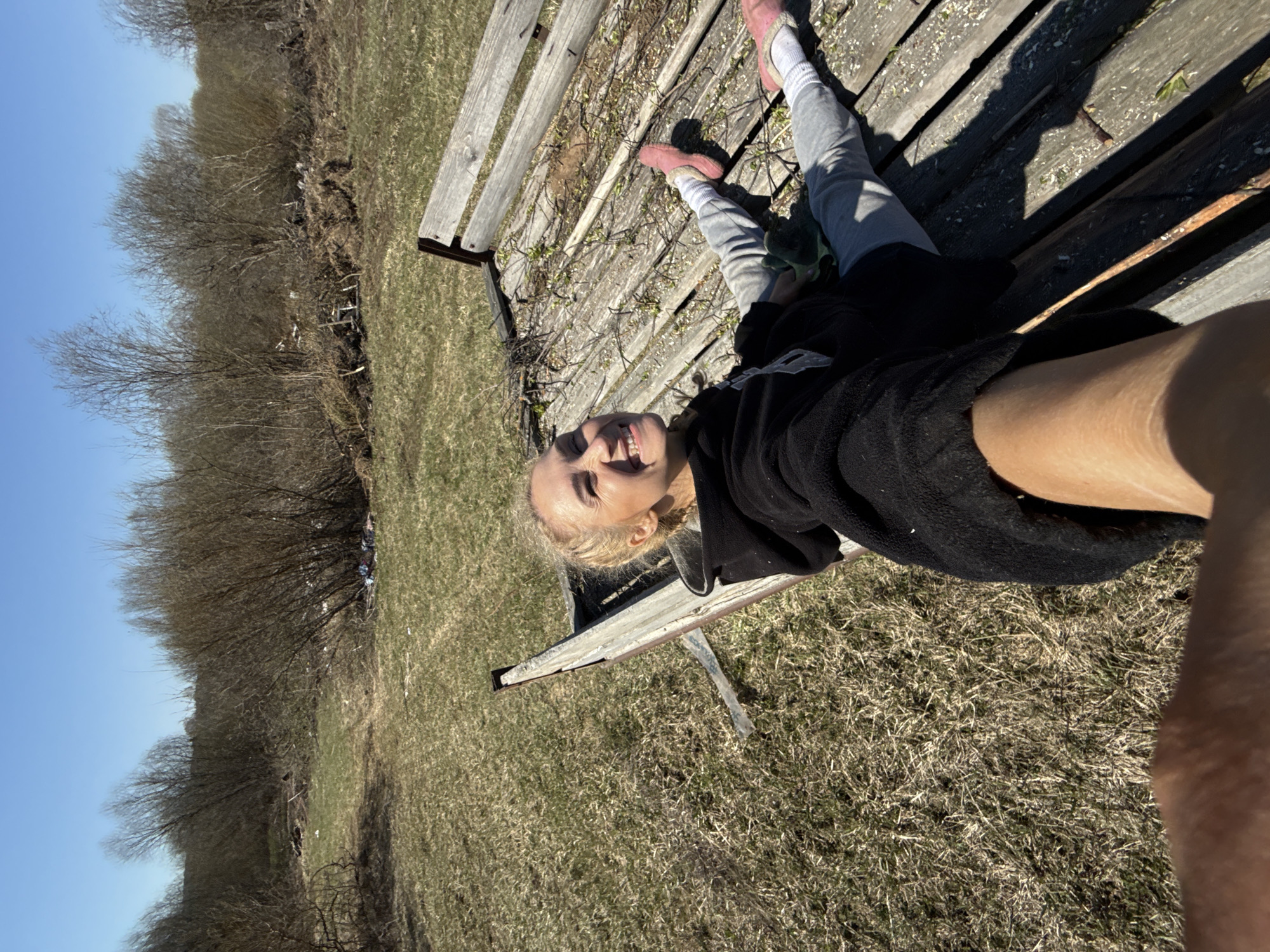 A young woman lies on a wooden bench in Nizhnyaya Chermoda, Russia, with her legs extended towards the upper right corner of the image. She wears gray leggings, white socks, and pink slip-on shoes. A black sweatshirt is draped over her upper body, and dark shorts cover part of her thighs. The woman has blonde hair pulled back and is smiling at the camera. The bench is weathered and positioned in a field of dry grass. In the background, there are bare trees against a clear blue sky.