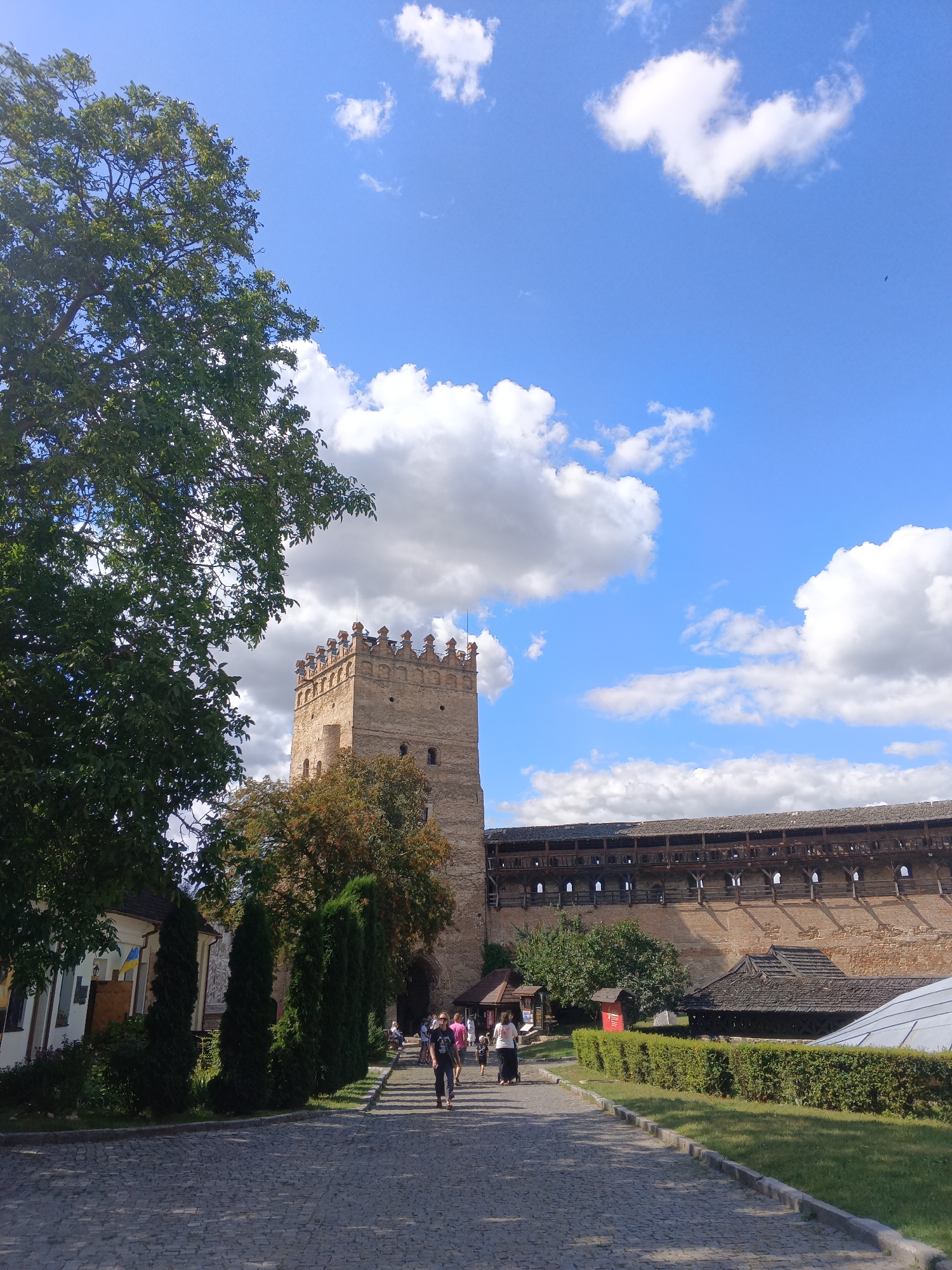 The image depicts a sunny day with a blue sky and white, fluffy clouds, likely taken in Lutsk, Ukraine. In the foreground, a cobblestone pathway leads towards a historic stone tower, the Lubart's Castle tower, which is the prominent structure in the image. The tower is made of light brown stone and has a crenellated top. To the right of the tower, a long, wooden-roofed wall with arched openings extends, forming part of the castle's fortifications.
A lush green lawn with neatly trimmed hedges borders the pathway on the right. On the left side of the pathway, there is a white building with a Ukrainian flag hanging outside and a large green tree with dense foliage. Several people are walking along the pathway towards the castle entrance. Some are dressed in casual summer attire. A group of people, including a woman pushing a stroller, are seen walking away from the camera. The overall scene conveys a sense of historical significance and tourist activity.