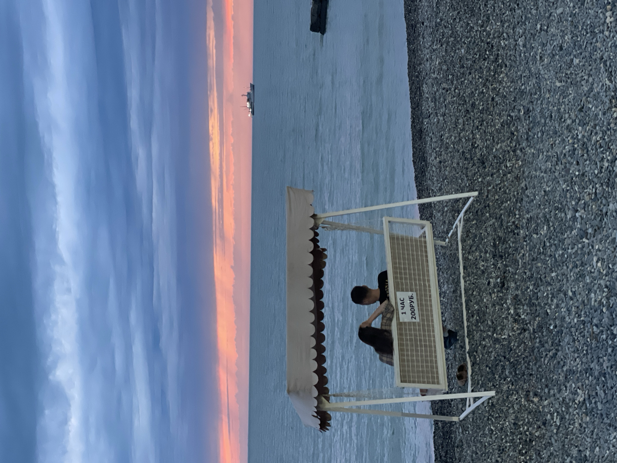 The image shows a pebble beach on the Black Sea with a view of a colorful sunset sky. In the background, a ship is visible on the horizon. In the foreground, there is a white metal bench with a canopy, where a man and a woman are sitting. A sign on the bench indicates a price of 200 rubles per hour. The location is most likely in the city of Sochi, Russia, based on the Russian script used on the sign.