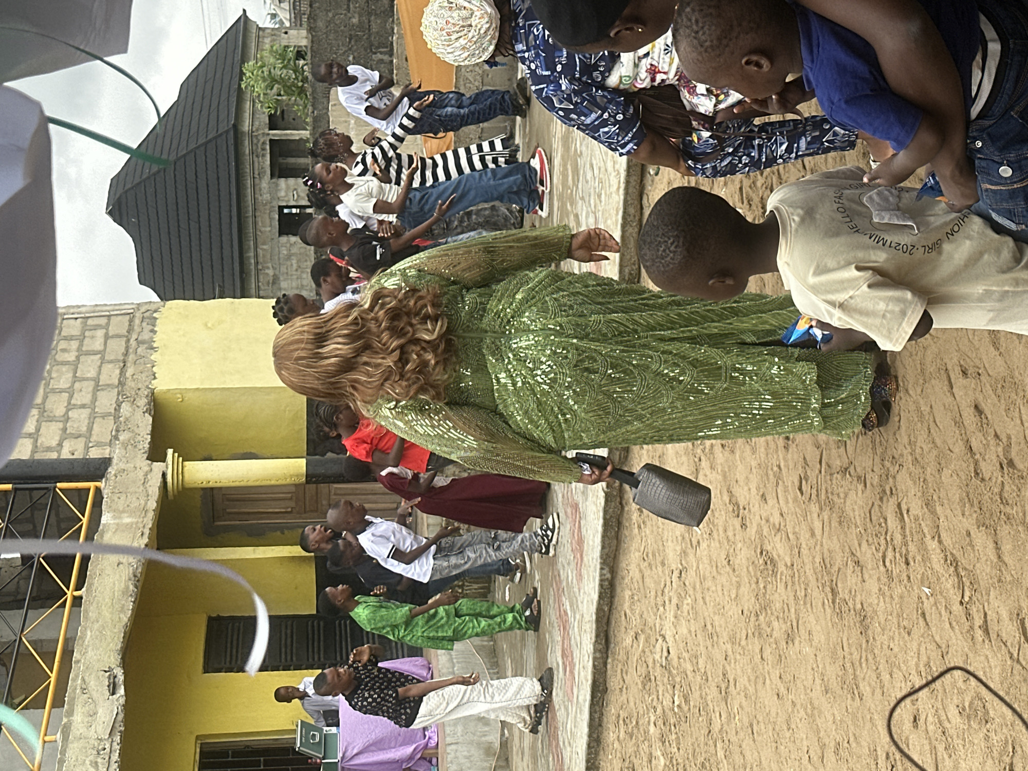 In Badagry, Nigeria, a group of children and adults are gathered outdoors in front of a building with a yellow facade and a gray cinder block wall. In the foreground, a woman in a sparkling green dress stands with her back to the camera, holding a dark-colored clutch bag. Several children are arranged in a line, facing away from the camera, some appearing to dance or engage in a performance. To the right, a woman in a blue and white patterned dress is seated, and a young boy in a blue shirt is on her lap. The ground is covered in sand. The background shows a portion of the building with a dark-tiled roof, possibly a residential structure. Decorative white and green ribbons are suspended from above. The overall scene suggests an outdoor event or gathering.