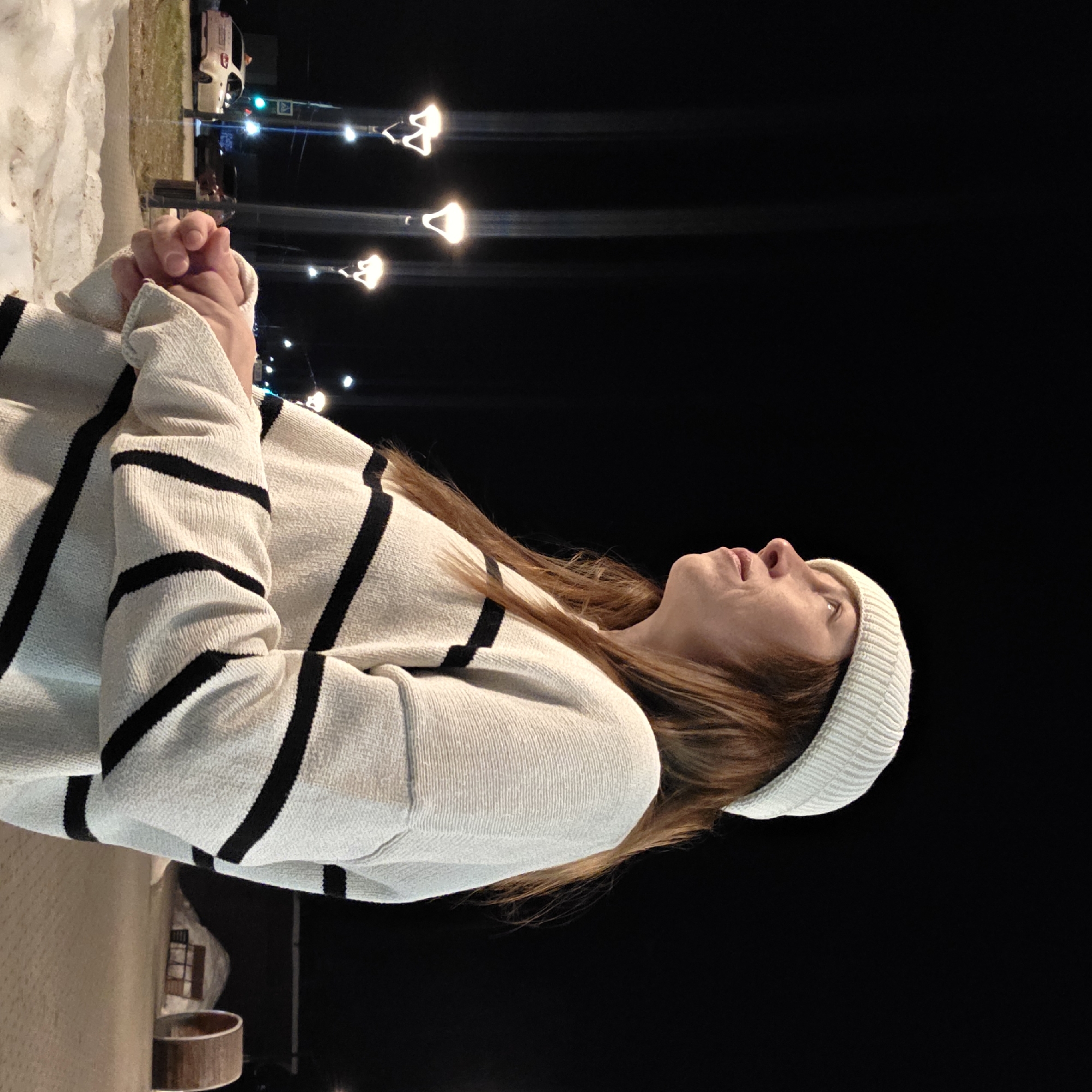 An outdoor nighttime scene in Chaikovsky, Russia, features a female subject positioned slightly left of center, viewed in profile from the mid-torso up, looking upwards and to the right. She wears a light beige knitted beanie and a light beige knitted sweater with thick black horizontal stripes. Her hands are clasped together in front of her. The immediate foreground shows patches of snow on the ground. The background on the left reveals a street illuminated by several streetlights with distinctive "W"-shaped fixtures, casting upward light trails. A white car, possibly a taxi, and a darker vehicle are visible on the street, alongside a green traffic light and a square road sign. The background on the right displays a paved area, a mound of snow, and a large, horizontally oriented wooden cylinder or barrel-like structure. A dark building is visible in the far background.