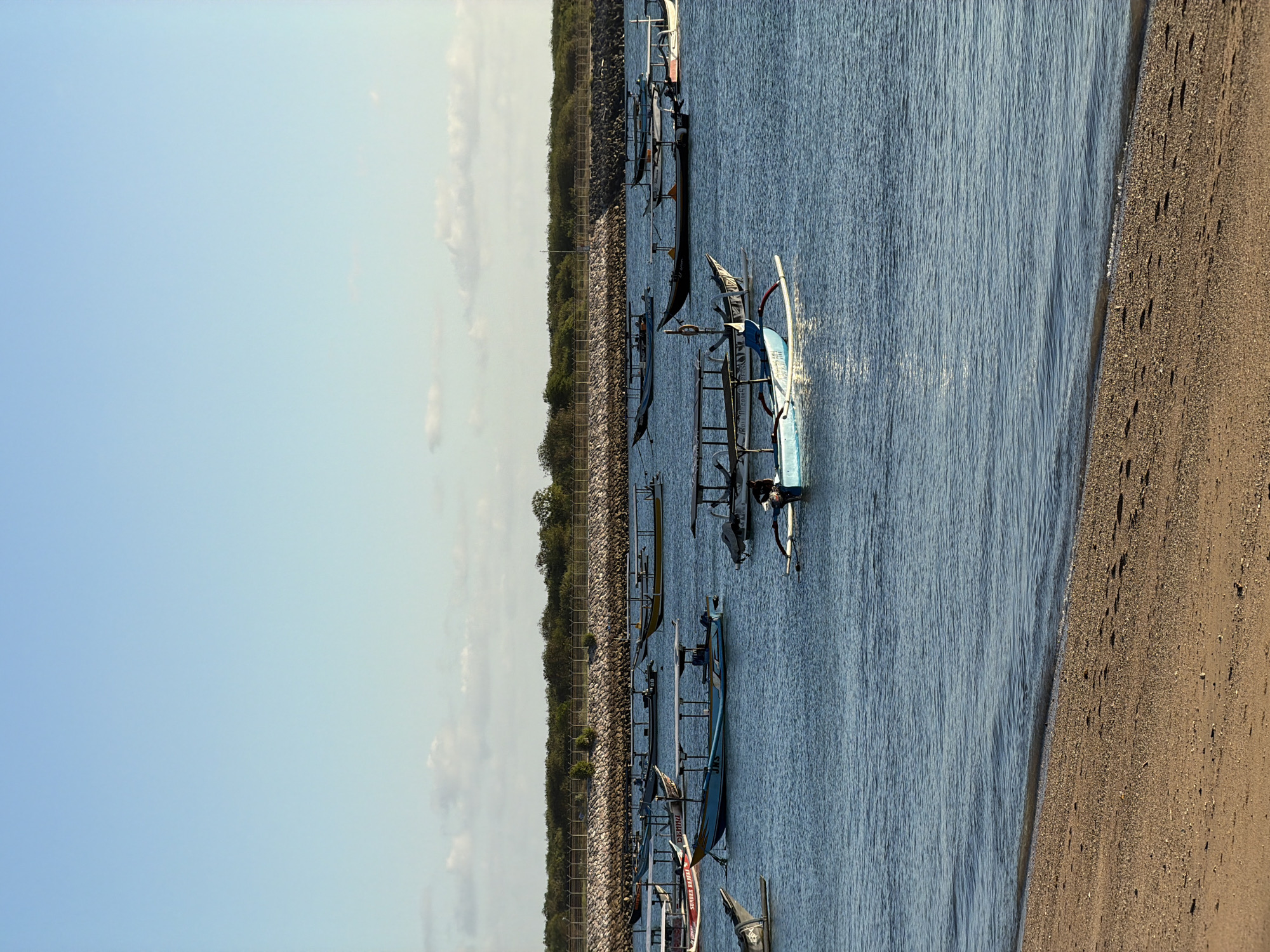 The image, correctly oriented, displays a serene coastal scene featuring numerous traditional Indonesian outrigger boats, known as jukungs, floating on calm, blue water. The sun, likely in the early morning or late afternoon, casts a warm glow and creates shimmering reflections across the water's surface.
In the foreground and middle ground, several jukungs are visible. A prominent light blue boat with white outriggers and a tall mast-like structure is positioned towards the center-right, appearing to be gently moving or recently docked, with small ripples around it. Text visible on this boat includes "BALI DEEP" and "BALI ADVENTURES". Other boats, some with colorful details like red and yellow, are scattered across the water, extending into the background. One boat has the number "59" painted on it, and another has partially visible text that appears to be "BALI OCEAN".
To the left of the boats, a rugged, rocky or concrete barrier lines the shore, providing a protective edge. Beyond this barrier, a band of lush green vegetation, possibly trees and bushes, stretches across the horizon. On the right side of the frame, a sandy beach slopes gently into the water, showing subtle ripples and traces of footprints, suggesting recent activity. The sky above is a clear, light blue with minimal, faint clouds near the horizon. No people are visible in the scene. The overall atmosphere is peaceful and tranquil, characteristic of a tropical coastal setting in Kuta, Indonesia.