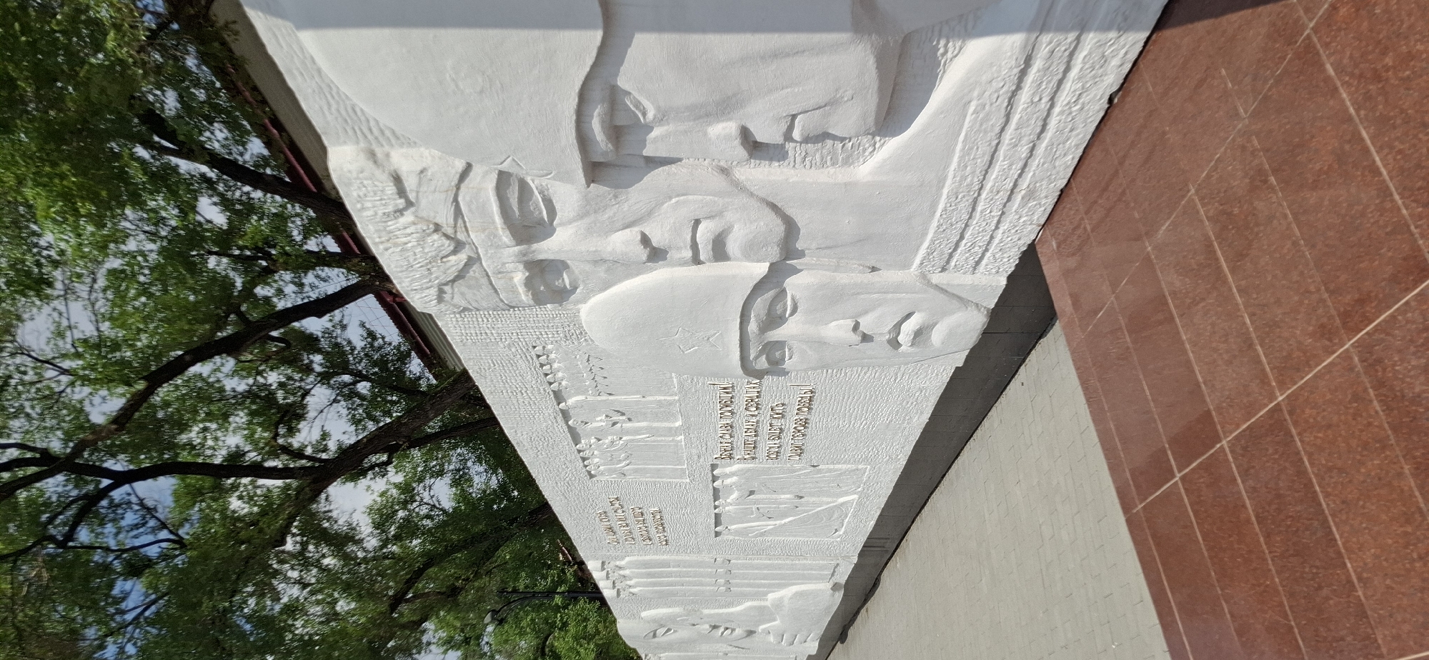 This image shows a large white bas-relief sculpture depicting soldiers. The sculpture is located in Blagoveshchensk, Russia, at the Eternal Flame monument. The monument is dedicated to soldiers who died in the Great Patriotic War. The sculpture depicts several soldiers' faces, some wearing helmets. The faces are rendered in a realistic style, with detailed features. The background of the sculpture features a procession of smaller figures, also in relief, representing soldiers marching. The sculpture is set against a white wall. In the foreground, there are red granite tiles and a paved walkway. Green trees with dark branches are visible above the wall, and a clear sky is visible through the leaves. The overall impression is one of solemnity and remembrance.