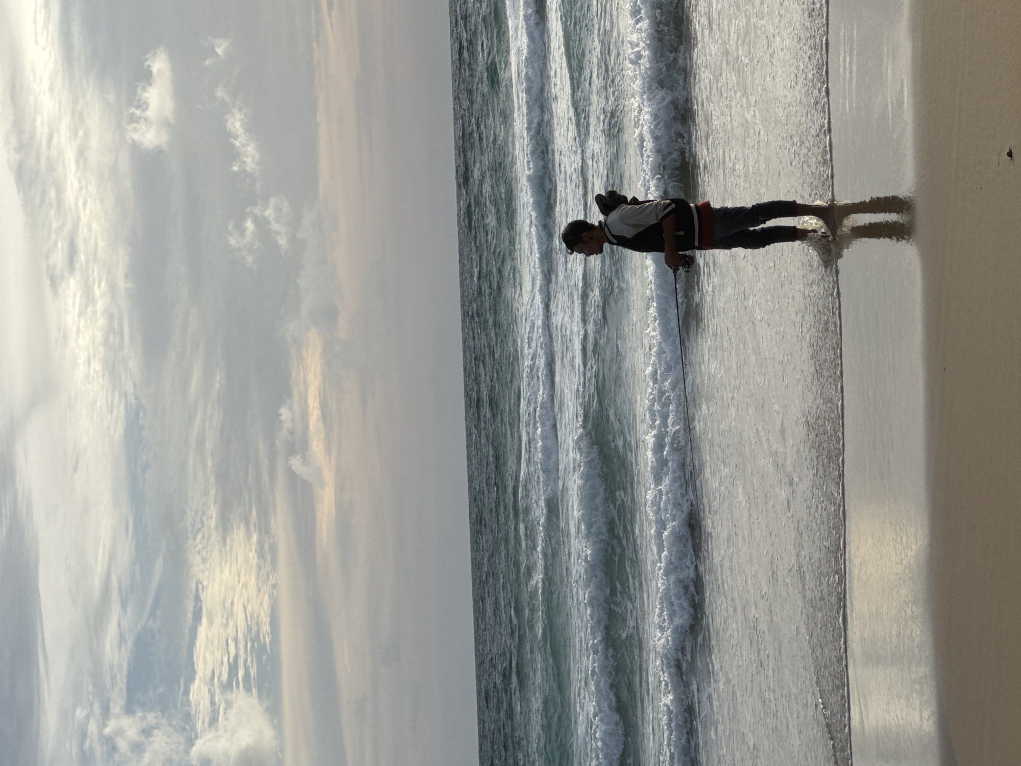 A lone figure stands in the shallow surf of Karon Beach, Thailand, fishing. The person, seen in profile, is wearing a dark jacket over a lighter shirt and dark pants. They hold a fishing rod and reel, casting into the choppy, turquoise waters. Waves crash onto the sandy shore, creating white foam that washes around the angler's bare feet. The sky above is a blend of soft grays and blues, with scattered clouds illuminated by the late afternoon sun. The overall scene conveys a sense of tranquility and solitude.