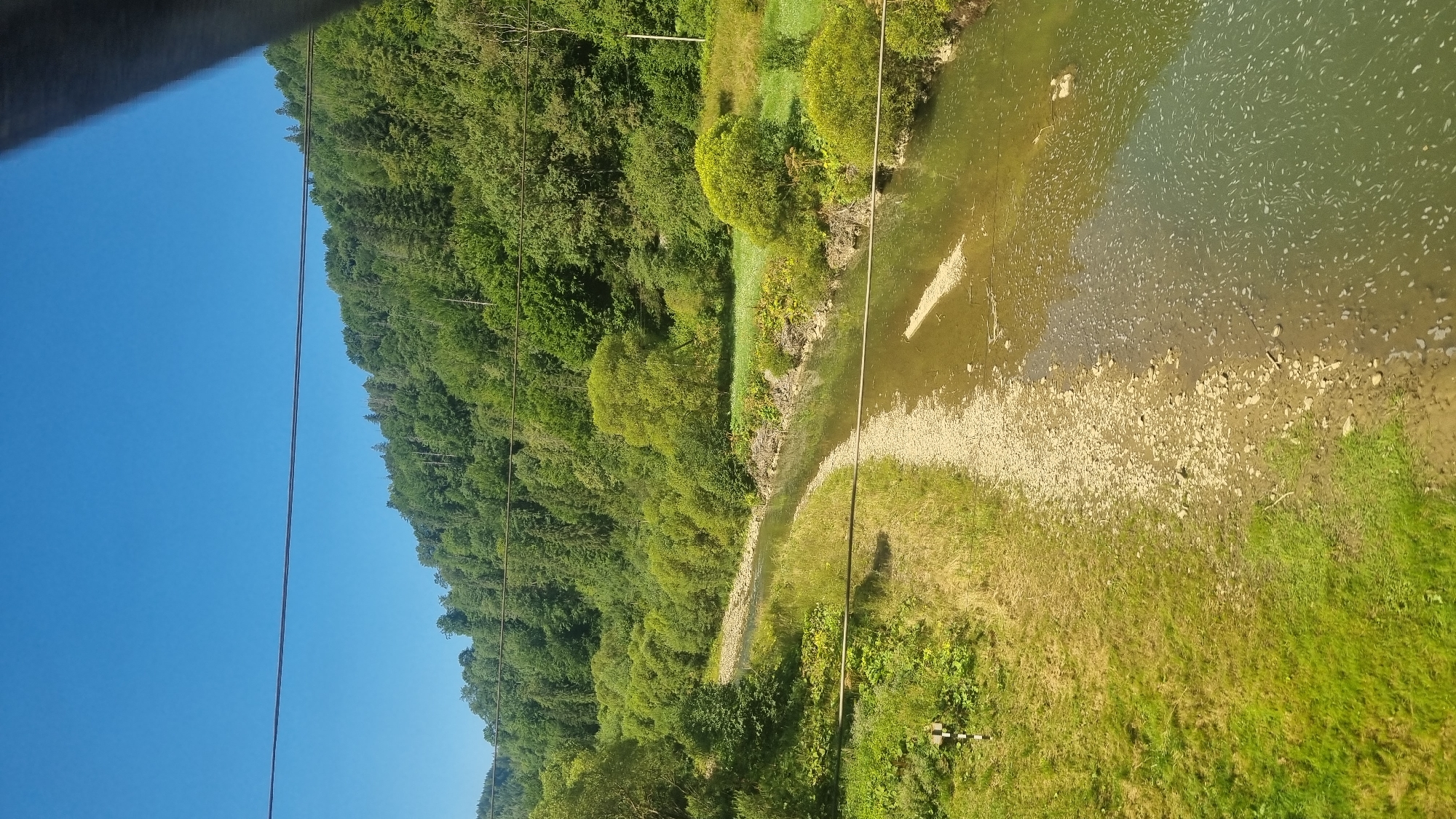 The image depicts a clear blue sky over a densely forested, hilly landscape. A winding river flows through the valley, with a rocky bank on one side and grassy slopes on the other. The river's water is a murky green, with some white foam visible on the surface, suggesting a slight current or turbulence. The trees on the hills are lush and green, indicating a summer or late spring setting. In the foreground, there are some power lines or cables stretching horizontally across the frame. The scene is tranquil and appears to be in a rural, natural environment. The location in Tukhlya, Ukraine, is a mountainous region known for its scenic beauty and natural landscapes, consistent with the image. There are no people or animals visible in the image.
