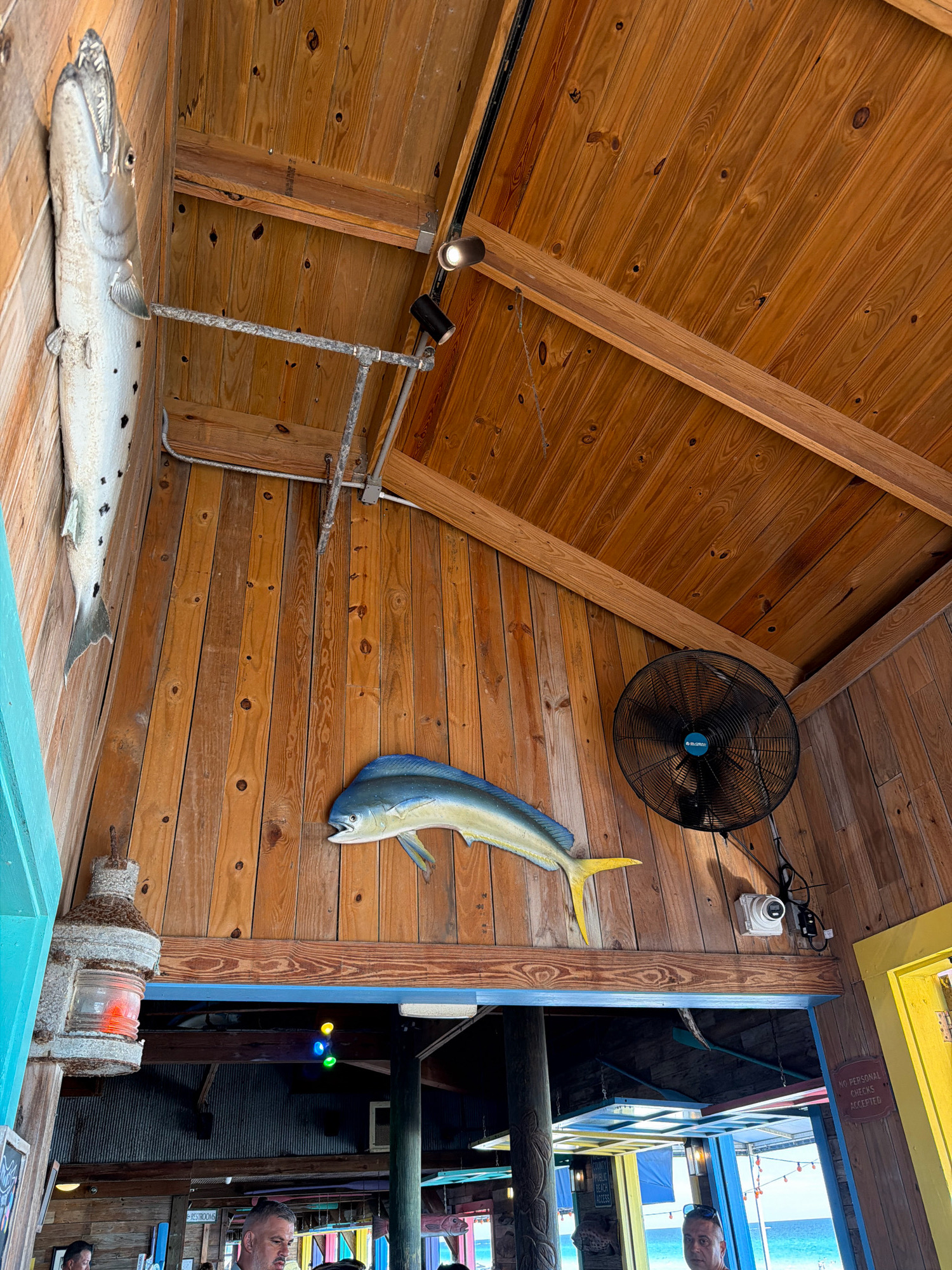 The image depicts the interior of a restaurant or bar with a rustic, nautical theme, likely located in Miramar Beach, Florida, United States of America. The walls and ceiling are constructed of warm-toned wood planks, creating a cozy and inviting atmosphere. Decorative elements include a large mounted fish on the left wall and a smaller, colorful mounted fish on the right wall. Above the entrance to another area, colored lights are visible. A black oscillating fan is mounted on the wall, and a security camera is positioned nearby. To the left, a vintage-style lantern with a red glass shade is attached to the wall. The lower portion of the image shows a glimpse of patrons and beachgoers, with a view of the ocean in the background, suggesting a beachfront location. The overall impression is a casual, possibly seafood-focused establishment.