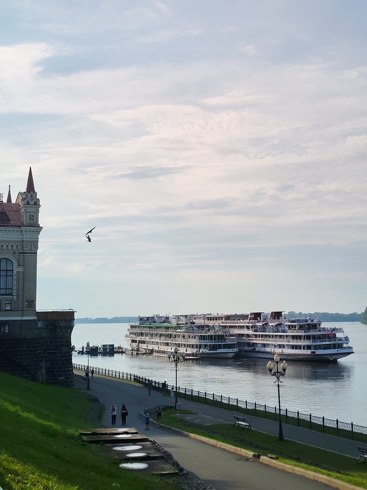 This media file shows a waterside scene in Bol'shie Mkhi, Russia. The setting is a riverfront promenade with a building on the left and large cruise ships docked on the right. The building on the left appears to be a historic structure with ornate architecture, featuring a peaked roof and multiple windows. It is situated on a grassy embankment.
In the foreground, a paved pathway runs along the river, bordered by a black metal fence and lampposts. Several people are visible on the promenade, walking and standing. A child is also present. The river is calm and reflects the sky.
Dominating the right side of the frame are at least two large, multi-decked cruise ships, docked side-by-side. They are white with red accents and appear to be passenger vessels. One ship has a Russian flag visible.
The sky is partly cloudy with a bright, hazy quality, suggesting it is daytime, likely late afternoon or early evening due to the soft lighting. The overall mood is peaceful and serene. No specific event is clearly indicated, but the scene depicts a common day at a waterfront location with leisure activities.
