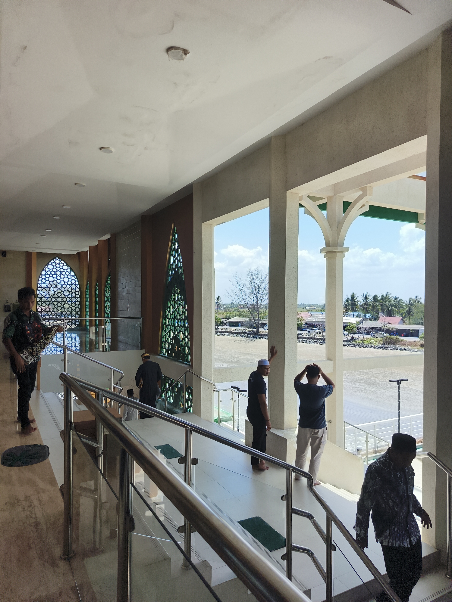 The image depicts the interior and exterior of a mosque in the city of Beringin, Indonesia. Inside, the architecture features white, textured ceilings with some water stains and circular light fixtures. A man in a patterned shirt and black pants stands on a polished floor, holding a prayer rug. A staircase with stainless steel railings and glass panels leads upwards, with several green prayer mats placed along the steps. Other men, some wearing traditional Islamic attire, are visible on the stairs and upper level. The interior is illuminated by natural light filtering through decorative patterned windows that are green in hue.
The mosque's exterior offers a view of a waterfront scene. A muddy or sandy shore is visible, with a body of water stretching towards a distant coastline. The coastline is lined with a mix of buildings, including houses with red and brown roofs, some shops, and scattered trees. Palm trees are prominent in the background, and the sky is blue with scattered white clouds. A few cars are parked near the buildings. The overall impression is of a coastal mosque in a tropical environment.