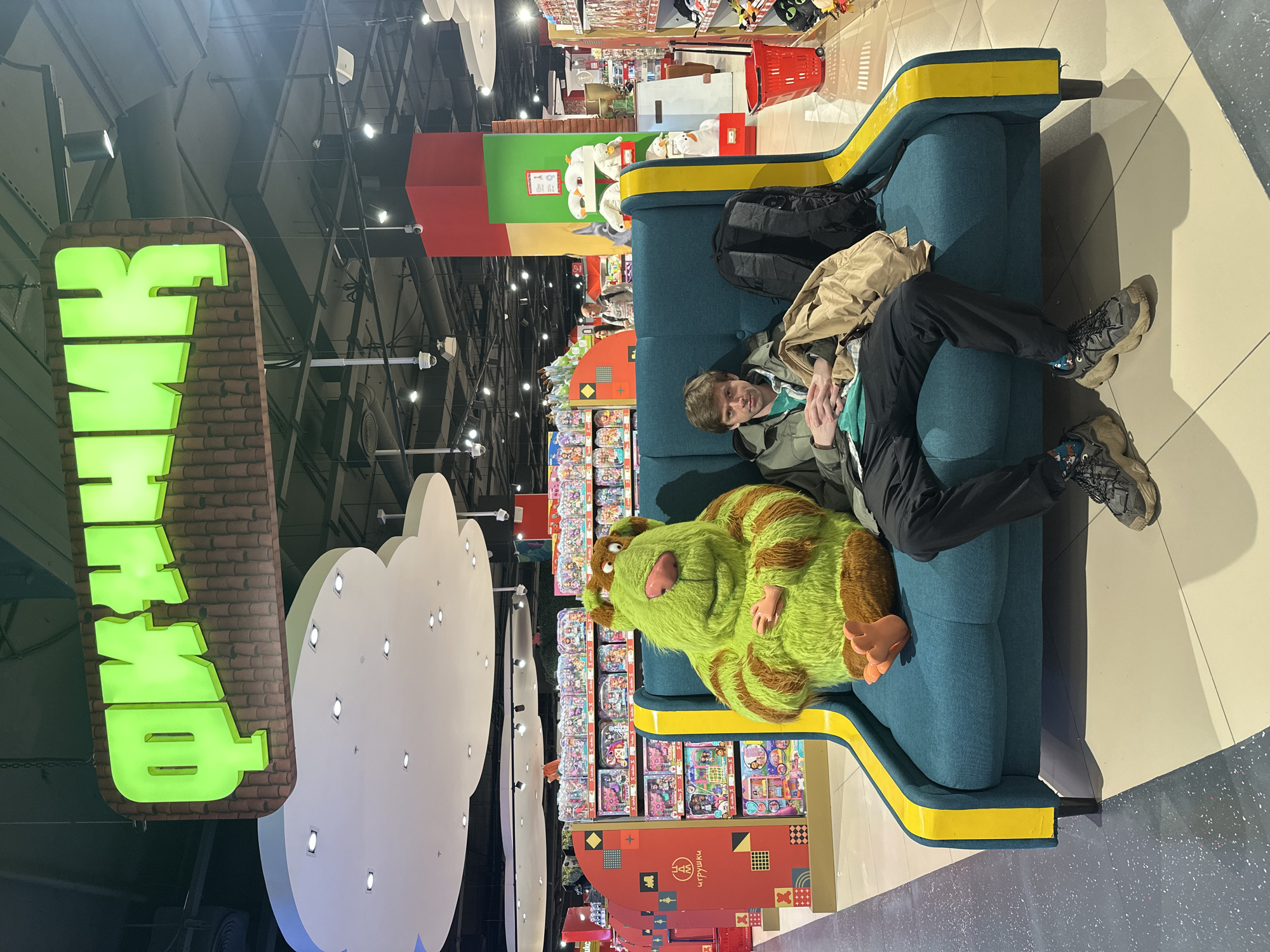 In a brightly lit indoor space, a man reclines on a blue couch with yellow trim, alongside a large, green and brown stuffed creature. The man wears dark pants, grey sneakers, and a green jacket, appearing relaxed. Behind the couch, shelves are stocked with colorful toy packaging. Overhead, a large, illuminated green sign with Cyrillic characters hangs adjacent to a cloud-shaped light fixture. Red shopping carts are visible in the background. This scene is located in a toy store in Moscow, Russia.