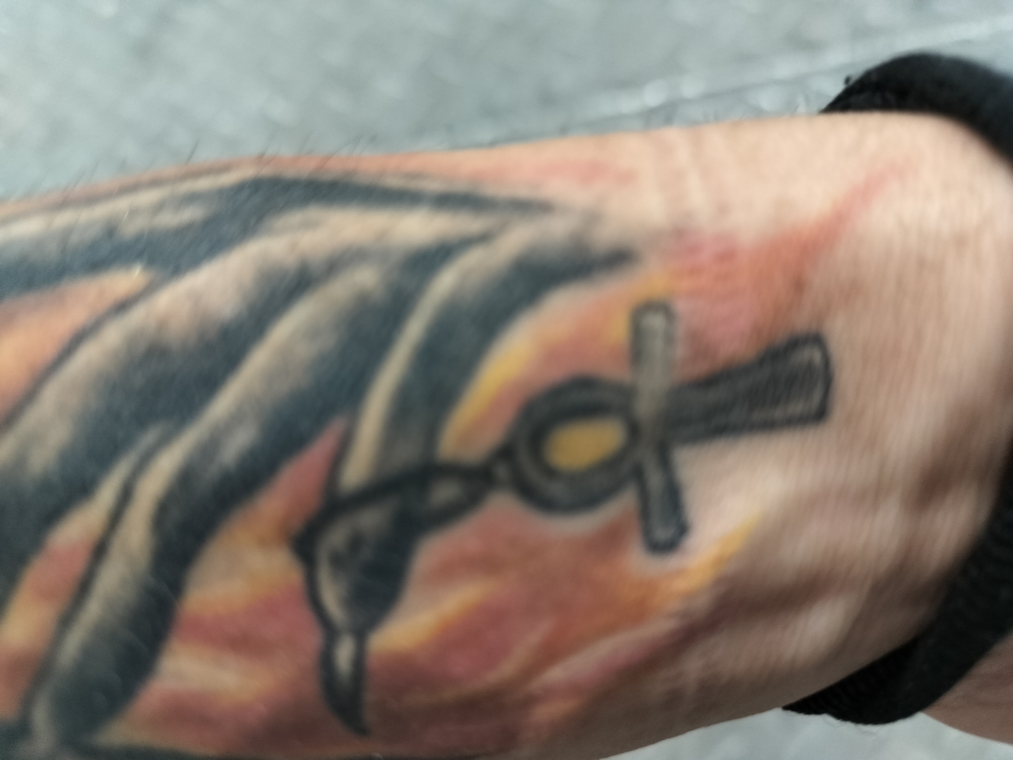 The image presents a close-up view of a person's forearm, prominently featuring a vibrant tattoo. The central element of the tattoo is an Ankh, an ancient Egyptian symbol of life, depicted with bold black outlines and a bright yellow-gold fill in its distinctive loop. Connected to the base of the Ankh is another dark-inked, stylized design, which appears to be a key or possibly an abstract creature, such as a bird's head or a serpentine form.
This central design is layered over a background of broader, dark, flowing tattoo strokes that span across the arm. Interspersed within and around these darker patterns are sections of warm red and orange ink, creating an effect reminiscent of fire, energy, or an abstract, colorful backdrop. The skin surrounding some parts of the tattoo shows a subtle redness, which might indicate recent tattooing or minor irritation.
On the wrist area, towards the right side of the image, a black elastic band or wristband is visible. The overall background is softly blurred and appears as a light grey or white surface, suggesting an indoor setting without specific environmental details. There is no discernible activity taking place as the arm is static for the photograph. No text is visible within the tattoo or elsewhere in the image. The time of day and weather are not ascertainable, and the provided location context of Gorno Sahrane, Bulgaria, is not visually supported by the image's content.