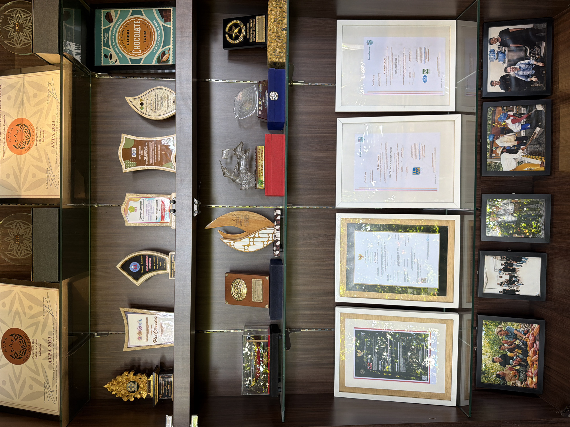 A well-lit, close-up shot shows a dark wooden display cabinet with three vertical glass-separated sections. The left section contains various award-like objects, including framed certificates, plaques, and golden trophies. Among them are boxes with "AVPA 2023" branding and a Lonely Planet "Global Chocolate Tour" box. The central section displays a similar collection of trophies and awards, with wooden and glass objects predominating. The right section houses framed certificates and black-framed photos of people, some engaged in cacao harvesting or processing activities. The displayed items suggest an Indonesian location specializing in chocolate, possibly in relation to awards for agricultural products.