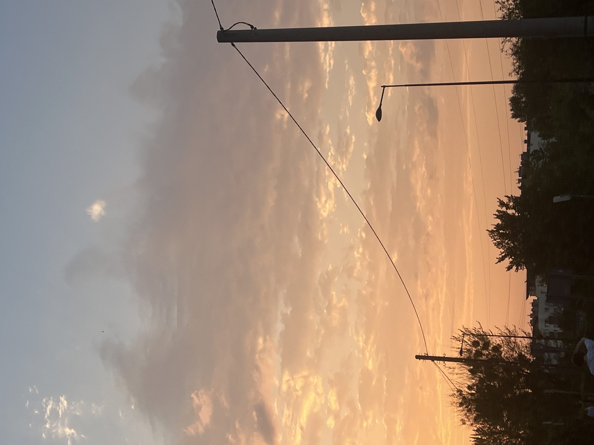 The image depicts a sunset scene with a sky filled with orange and pink clouds. In the foreground, there are two lampposts and some trees. In the background, there are buildings and more trees. The sun is setting, casting a warm glow over the scene. The overall mood is peaceful and serene. The image is taken in Warsaw, Poland.