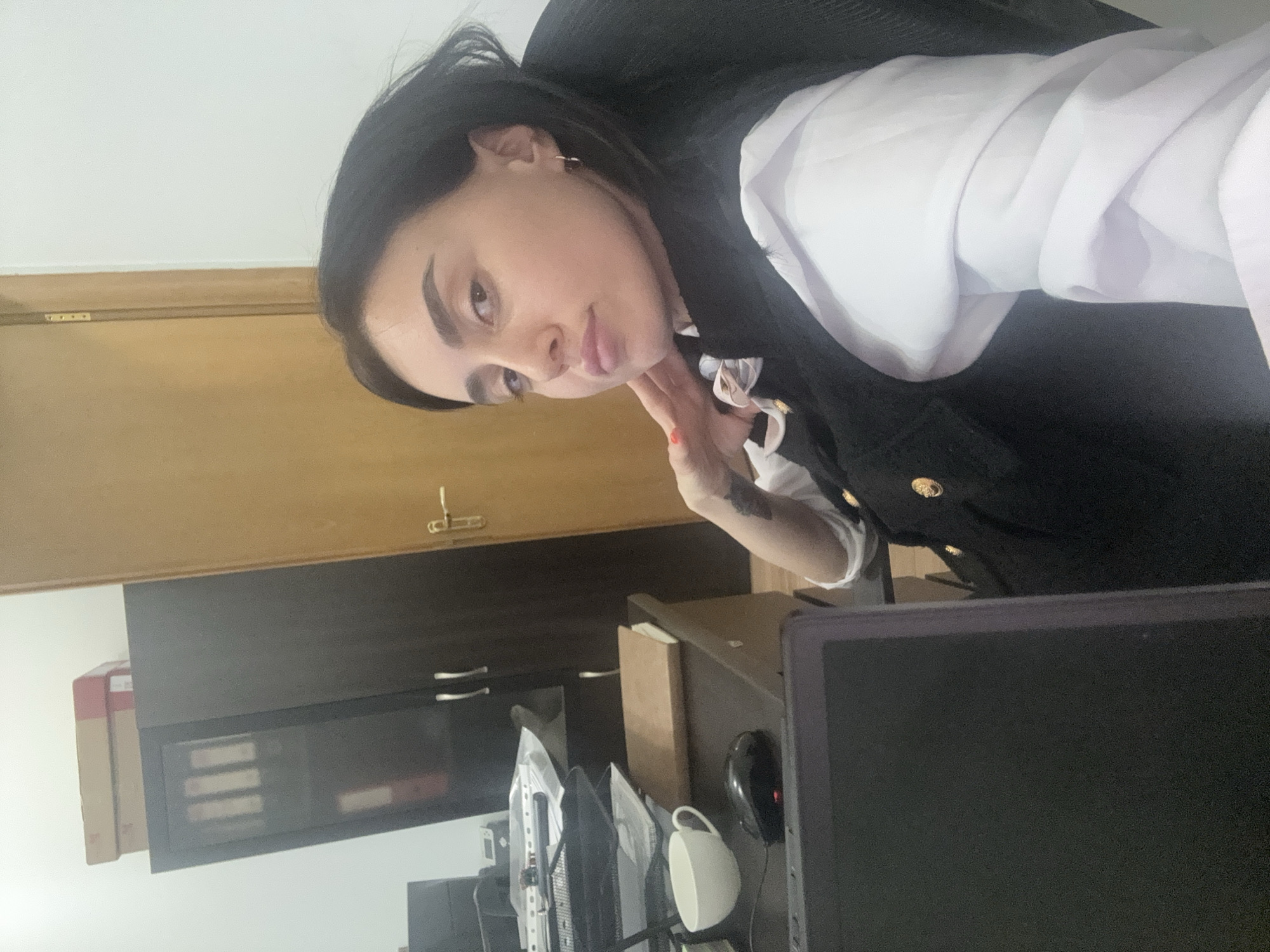 A portrait-style photograph, taken from a slightly low angle, shows a young woman looking directly at the viewer. She has dark hair, styled straight, and prominent dark eyebrows. Her eyes appear light-colored, and she has a subtle pout on her lips, conveying a pensive or slightly alluring expression. She is wearing a white collared blouse under a dark vest or jacket with gold buttons. Her right hand, with painted fingernails (possibly orange or red), is gently placed near her chin and neck. A tattoo is visible on her left forearm.
The setting appears to be an indoor office or workspace. Behind the woman, to her left, a light-colored wooden door or paneling is visible. To her right and in the background, a dark cabinet with two silver handles and a glass-fronted section is present, revealing some items inside.
In the foreground, a desk area is cluttered with office items. Several dark-screened computer monitors are visible, suggesting they are either off or displaying dark content. A white mug with a handle and a black computer mouse are on the desk. Other items include stacks of documents or folders in organizational trays, and a few cardboard boxes, one with red and white markings, are placed on and around the desk. The overall lighting suggests daytime. There is no discernible text visible in the image. The image's location context is Tbilisi, Georgia.