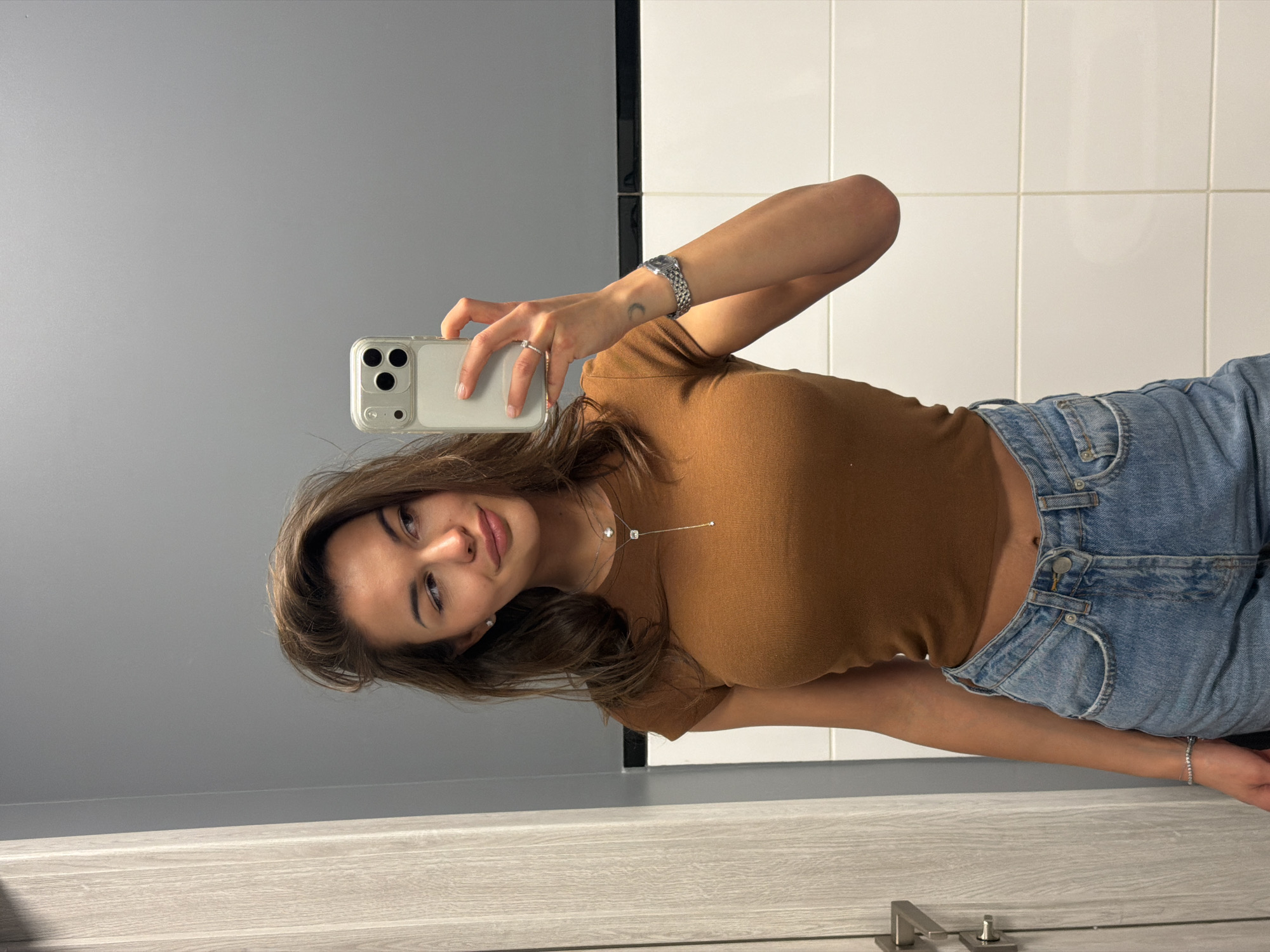 A young woman takes a mirror selfie, captured from a rotated perspective. She has brown hair, dark eyes, and a gentle smile, looking directly towards the camera. In her right hand, she holds a light-colored smartphone with a clear case and a triple-lens camera setup.
She wears a form-fitting, short-sleeved, brown crop top and light blue high-waisted denim jeans. Her left wrist features a silver watch with a metal band, and a silver ring is on her left ring finger. A delicate silver bracelet adorns her right wrist, while a thin silver necklace with a small square pendant is visible around her neck. Small stud earrings complete her look.
The indoor setting suggests a modern bathroom or changing room, typical of a location like Krasnoyarsk, Russia. The background is split by the mirror's reflection: white tiled walls with dark grout lines are on her left (viewer's right), and a plain grey wall is on her right (viewer's left). A light wood-grain surface, possibly a floor or countertop, is in the foreground, and a silver door handle is visible at the bottom right. The scene is brightly lit, likely by artificial light. There is no visible text in the image.