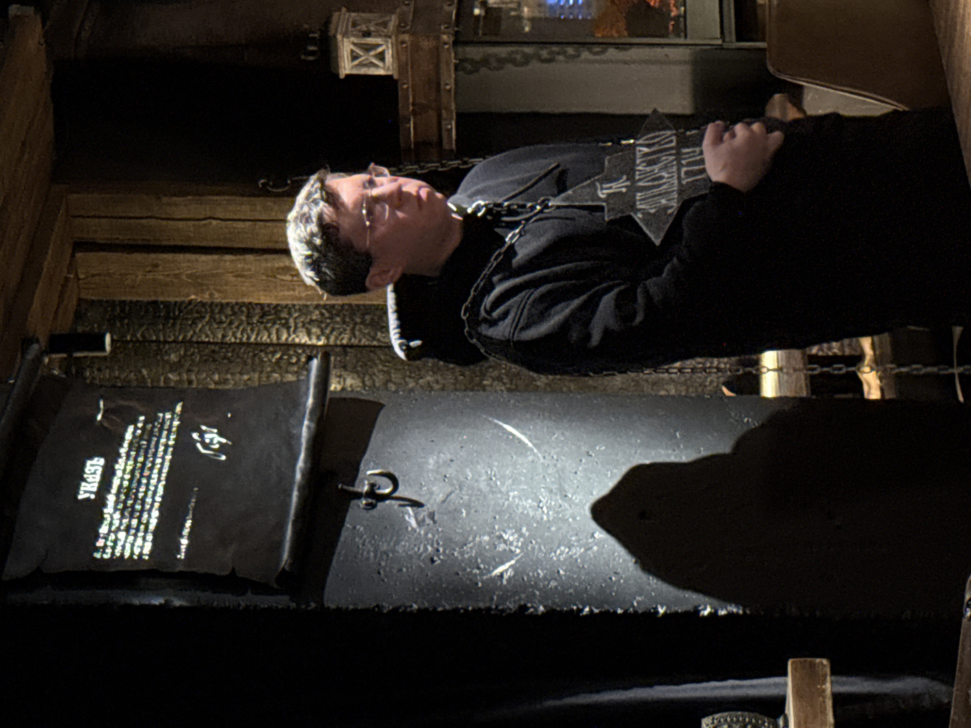 A Caucasian man is lying within an enclosed structure resembling a coffin or exhibit in what appears to be a themed attraction. He is wearing glasses and a dark-colored hooded sweatshirt, and a chain is draped around his neck. He is holding a sign with Cyrillic text. To the left of him, a scroll-like display with illuminated text in Russian is visible. The environment seems to be dimly lit and constructed with wood and metal, giving off a theatrical or historical display vibe. Given the mention of Kazan, Russia, this might be part of a historical or cultural exhibit or attraction within the city. The exact location and nature of the attraction can not be determined with the content provided.