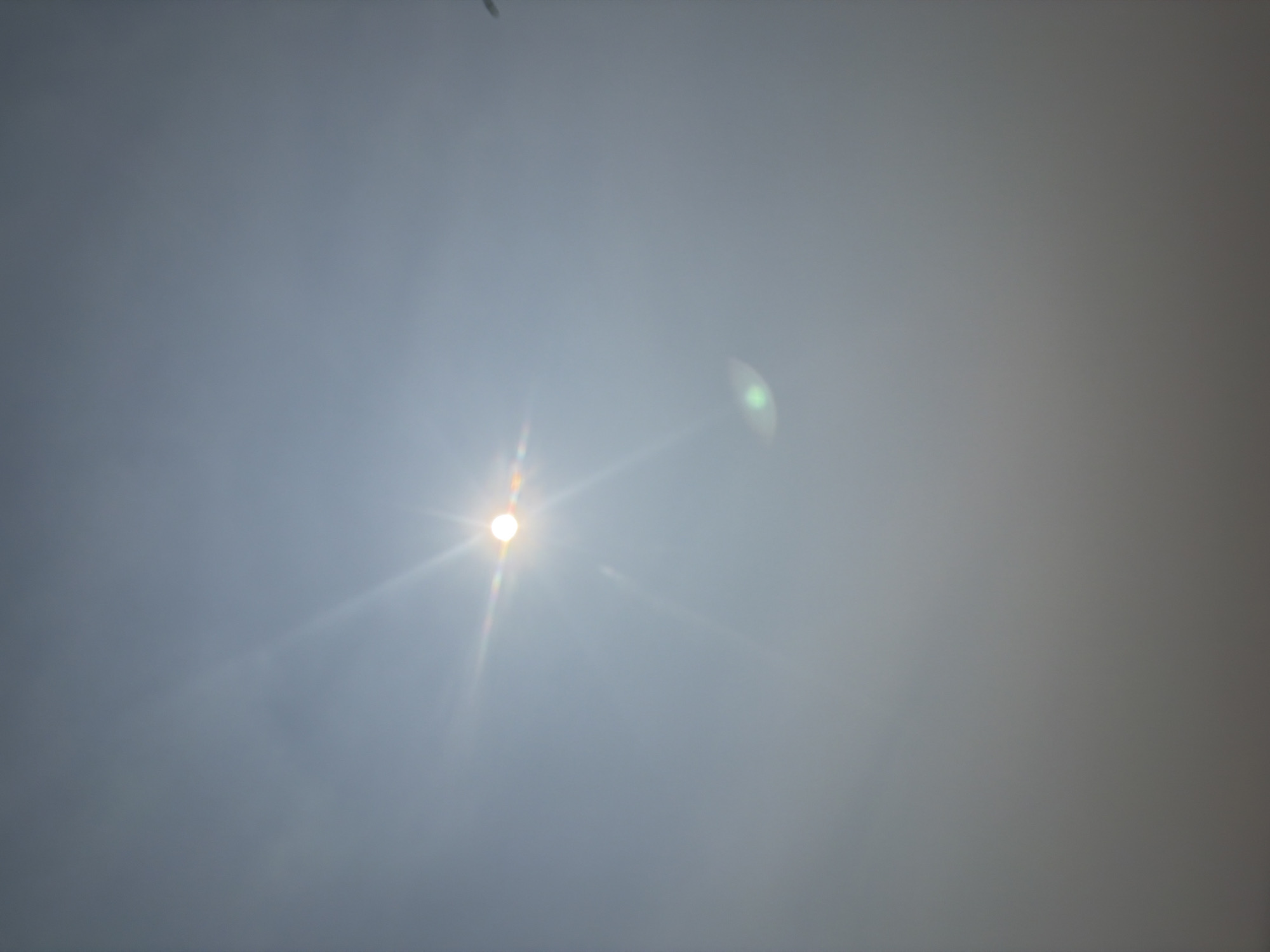A bright sun is visible in the upper center of the frame, casting lens flares and visible light rays across a hazy, light gray sky. A smaller, green lens flare is located to the lower right of the sun. The bottom portion of the image is obscured by a dense, gray haze, making it impossible to discern any details of the environment or ground. The location is identified as Myandigi, Russia. There are no people or specific objects clearly visible.